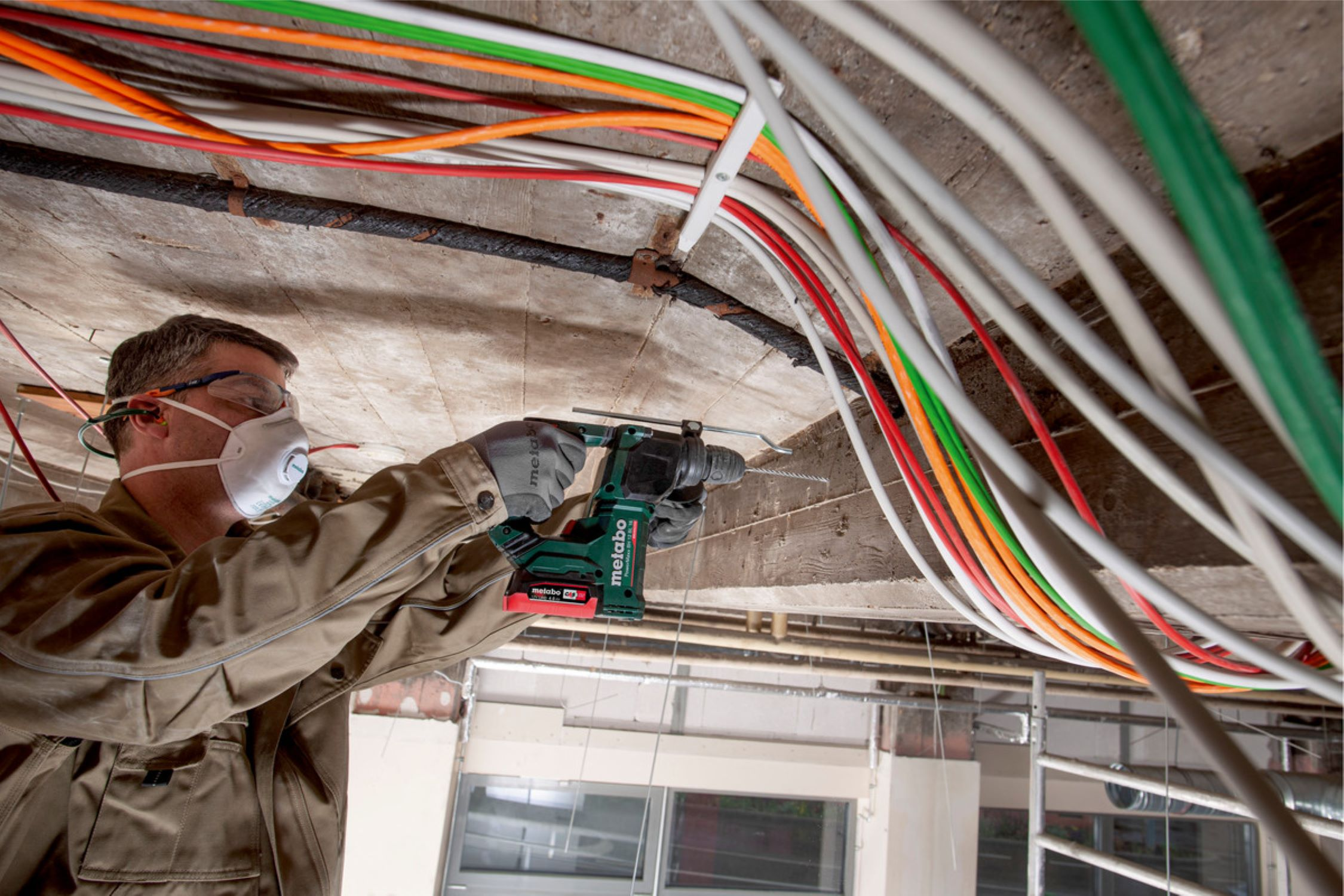 Ein Arbeiter bohrt in eine Betondecke inmitten eines Netzwerks farbiger Elektrokabel.