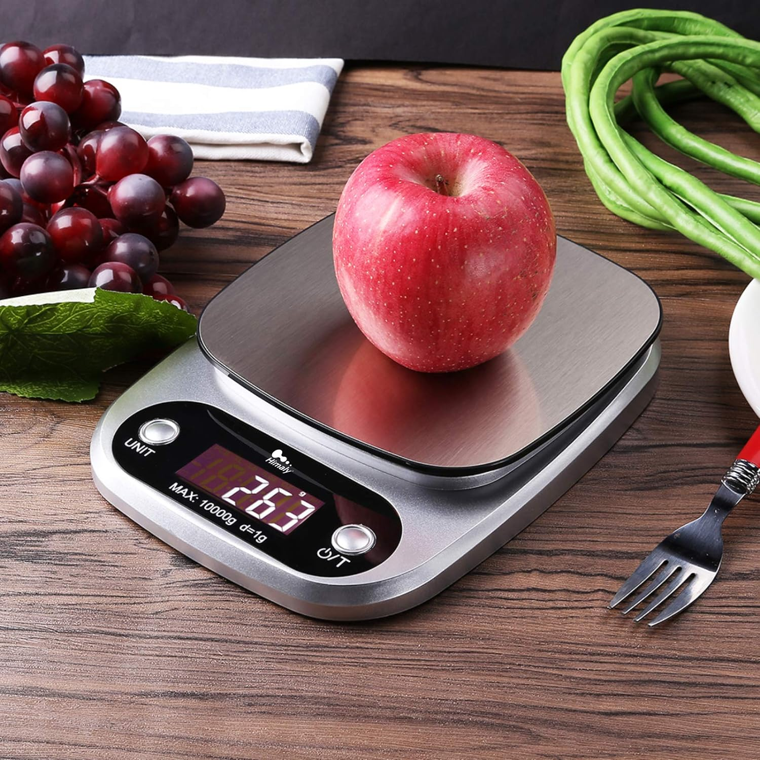 Digitale Küchenwaage mit Apfel. Anzeige zeigt Gewicht. Trauben, Gabel und Bohnen in der Nähe.