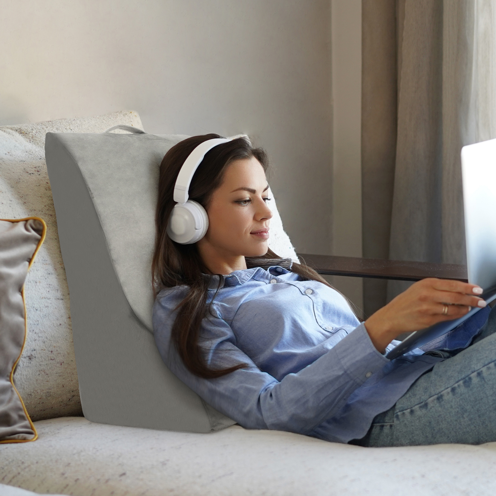 Frau benutzt Laptop, auf einem grauen Keilkissen ruhend. Sie trägt Kopfhörer und sitzt auf einem Sofa.