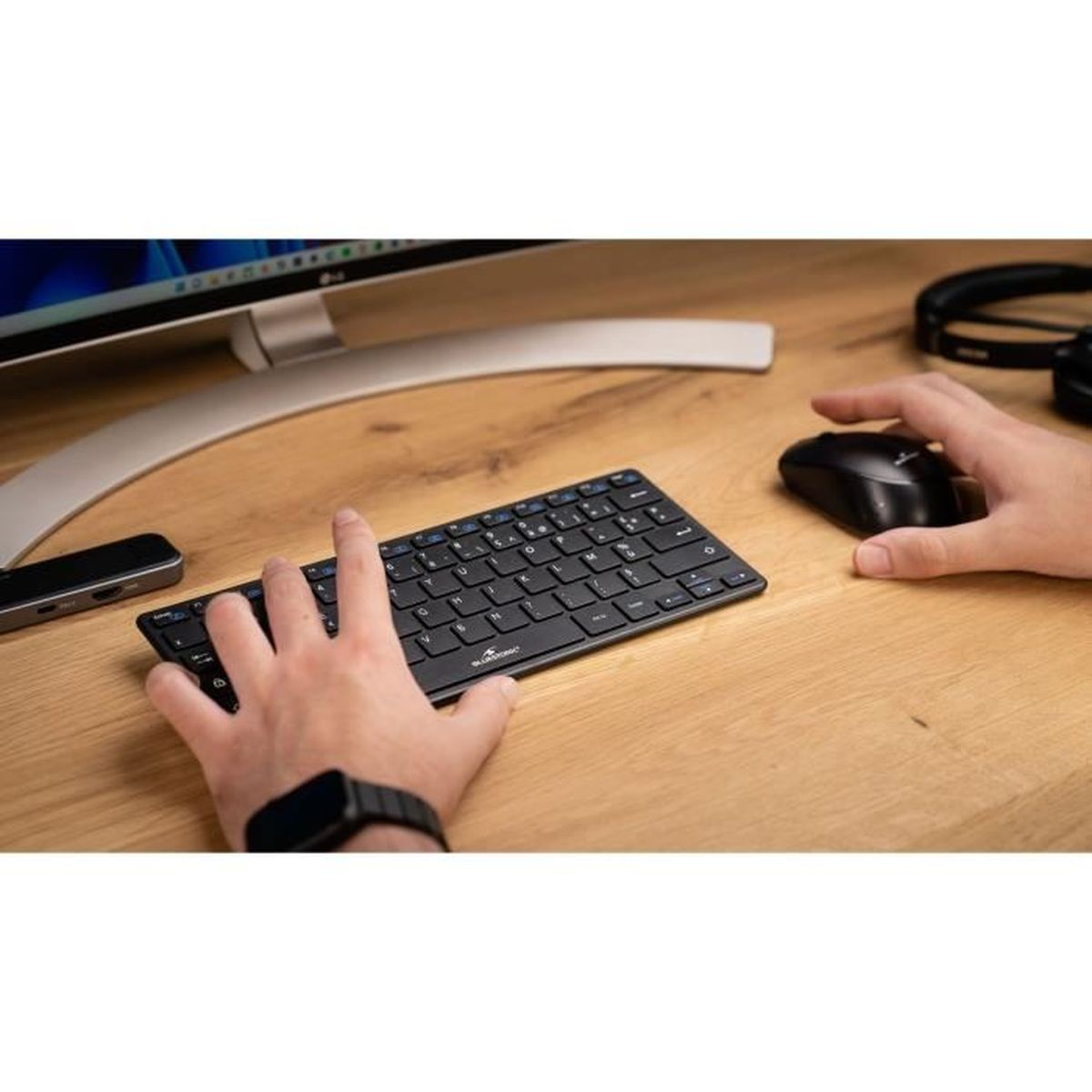 Hände benutzen schwarze Tastatur und Maus auf Holztisch. Monitor und Kopfhörer sichtbar.