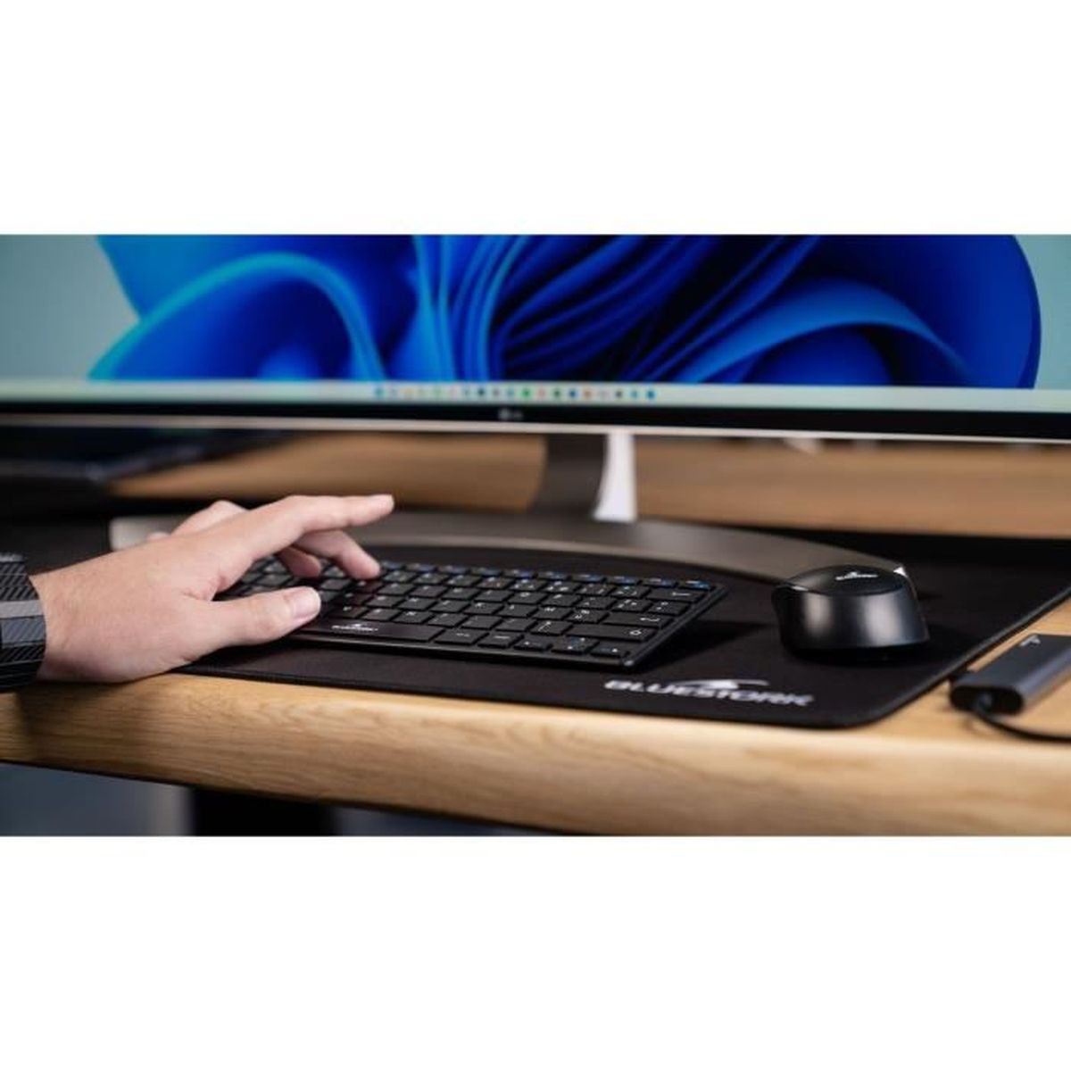 Person benutzt eine schwarze Tastatur und Maus auf einem Schreibtisch mit Monitor im Hintergrund.