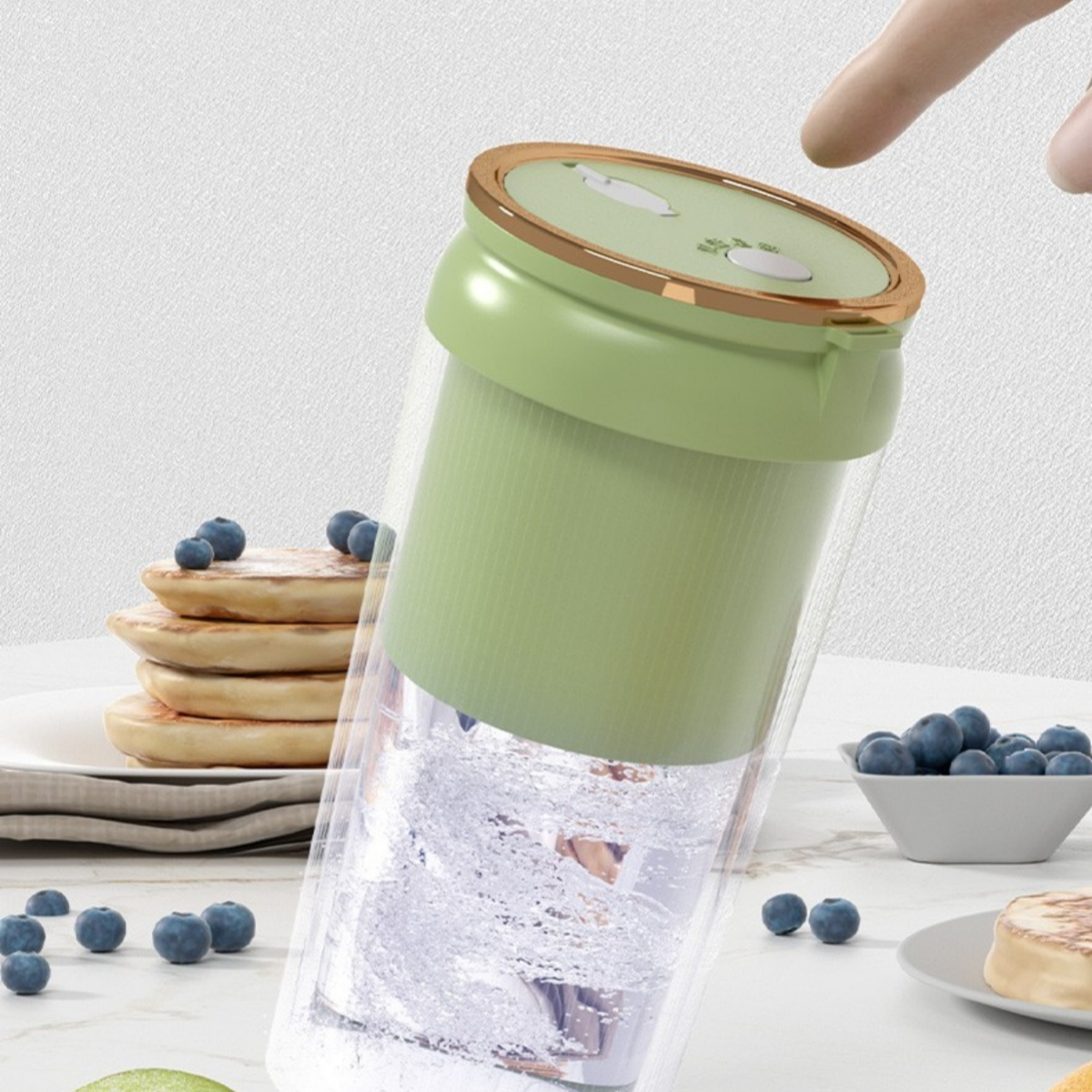 Grüner und goldener Mixer mit Pfannkuchen und Blaubeeren. Hand zeigt auf den Deckel.