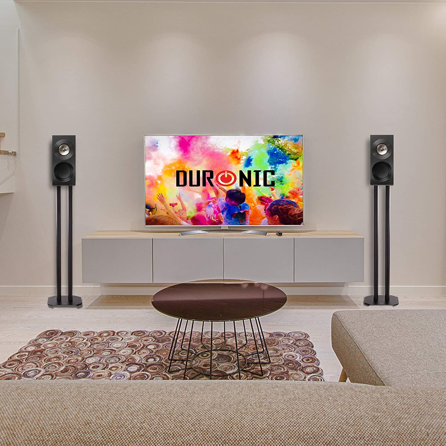 Wohnzimmer mit Fernseher, Lautsprechern, Couchtisch und Teppich. Der Fernseher zeigt ein farbenfrohes Bild.
