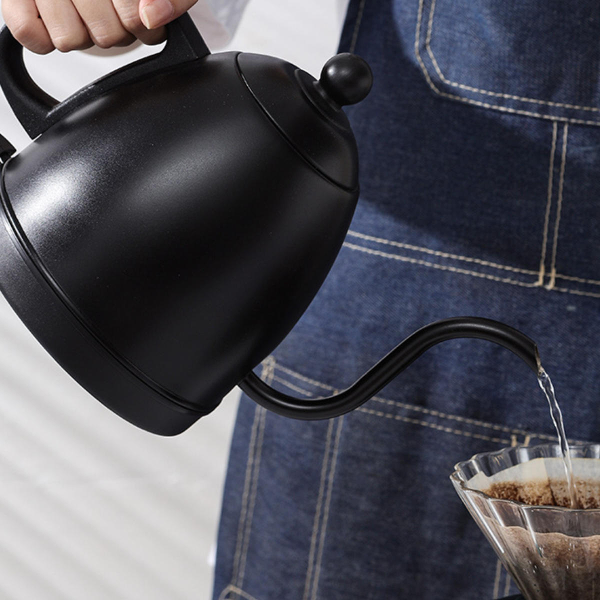 Ein schwarzer Schwanenhalskessel gießt Wasser in einen Glas-Pour-Over-Kaffeezubereiter, der von einer Person gehalten wird.