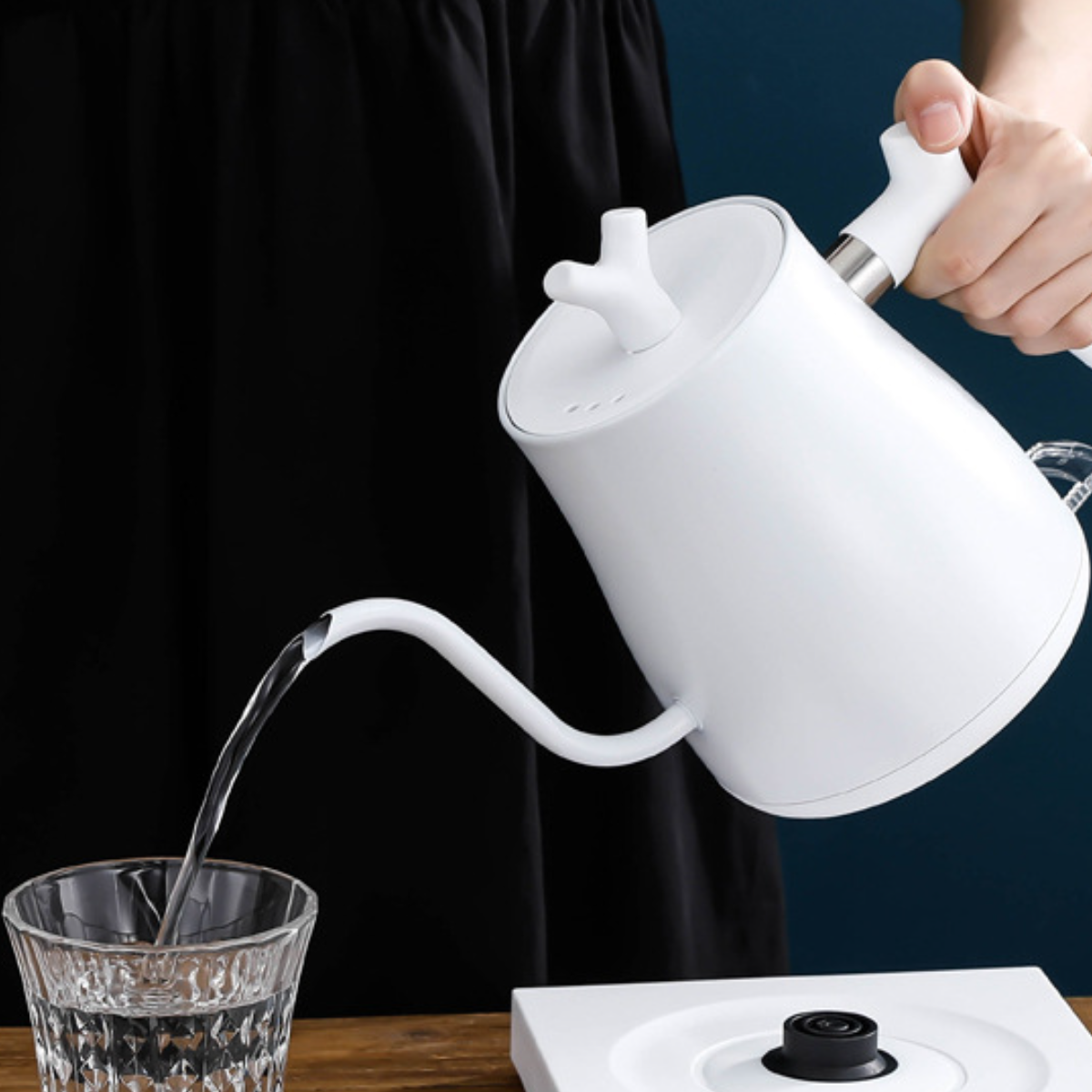 Weißer Wasserkocher gießt Wasser in ein Glas. Schwarzer Sockel, weißer Griff und Ausguss.