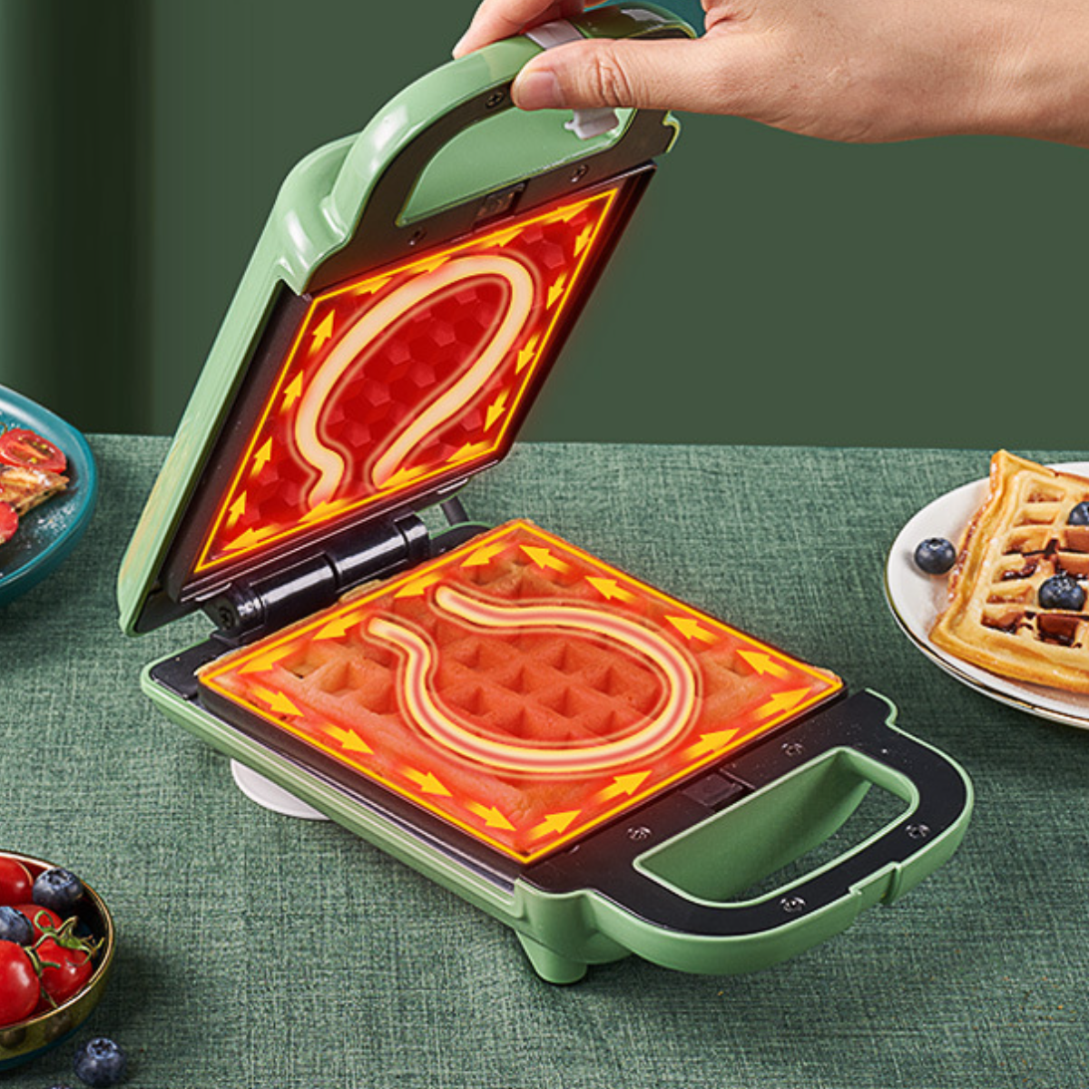 Grüner Waffeleisen offen, Waffeln darin, und eine Hand öffnet es. Waffel- und Beerenplatte auf dem Tisch.