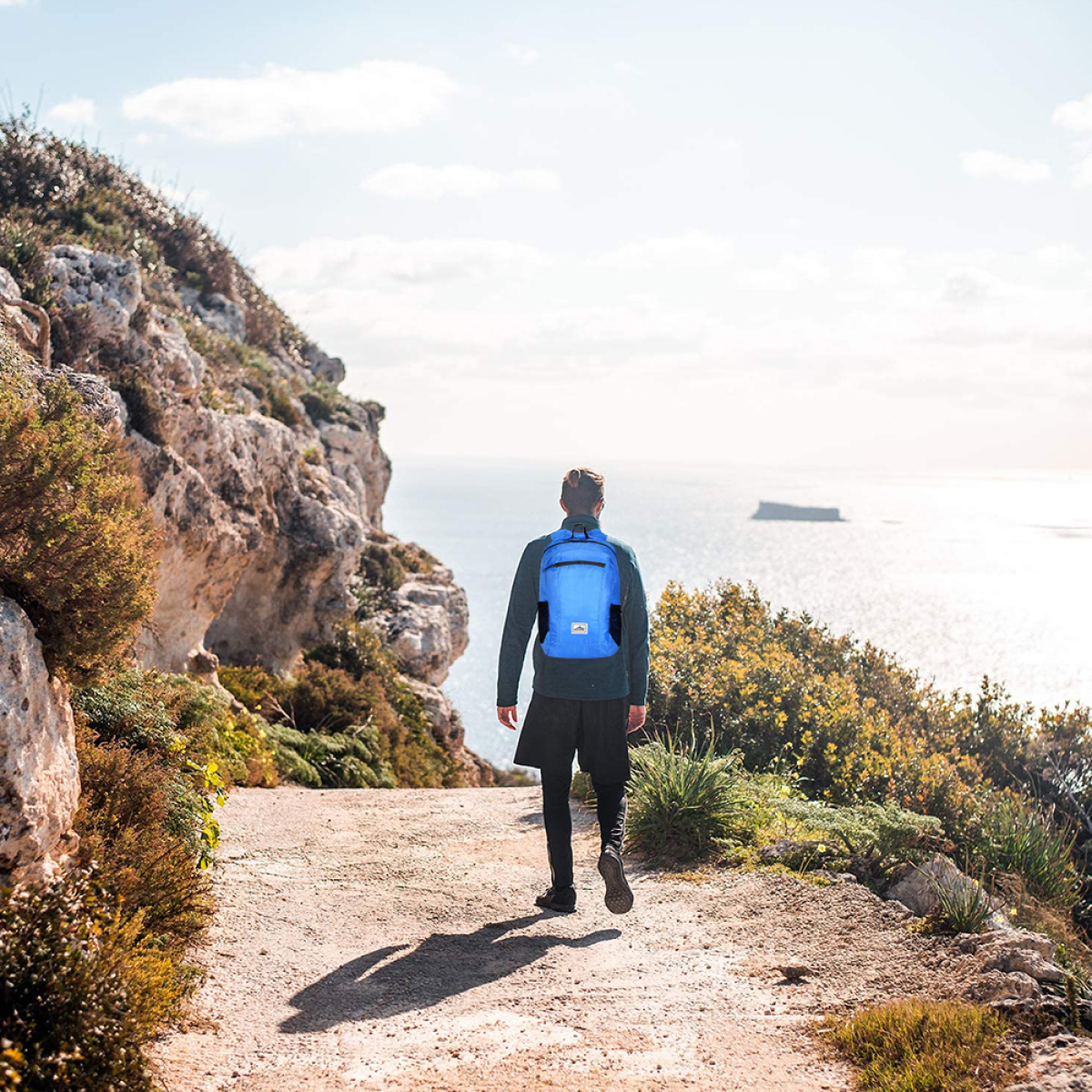 Eine Person, die auf einem Pfad zum Meer weggeht und einen blauen Rucksack trägt.