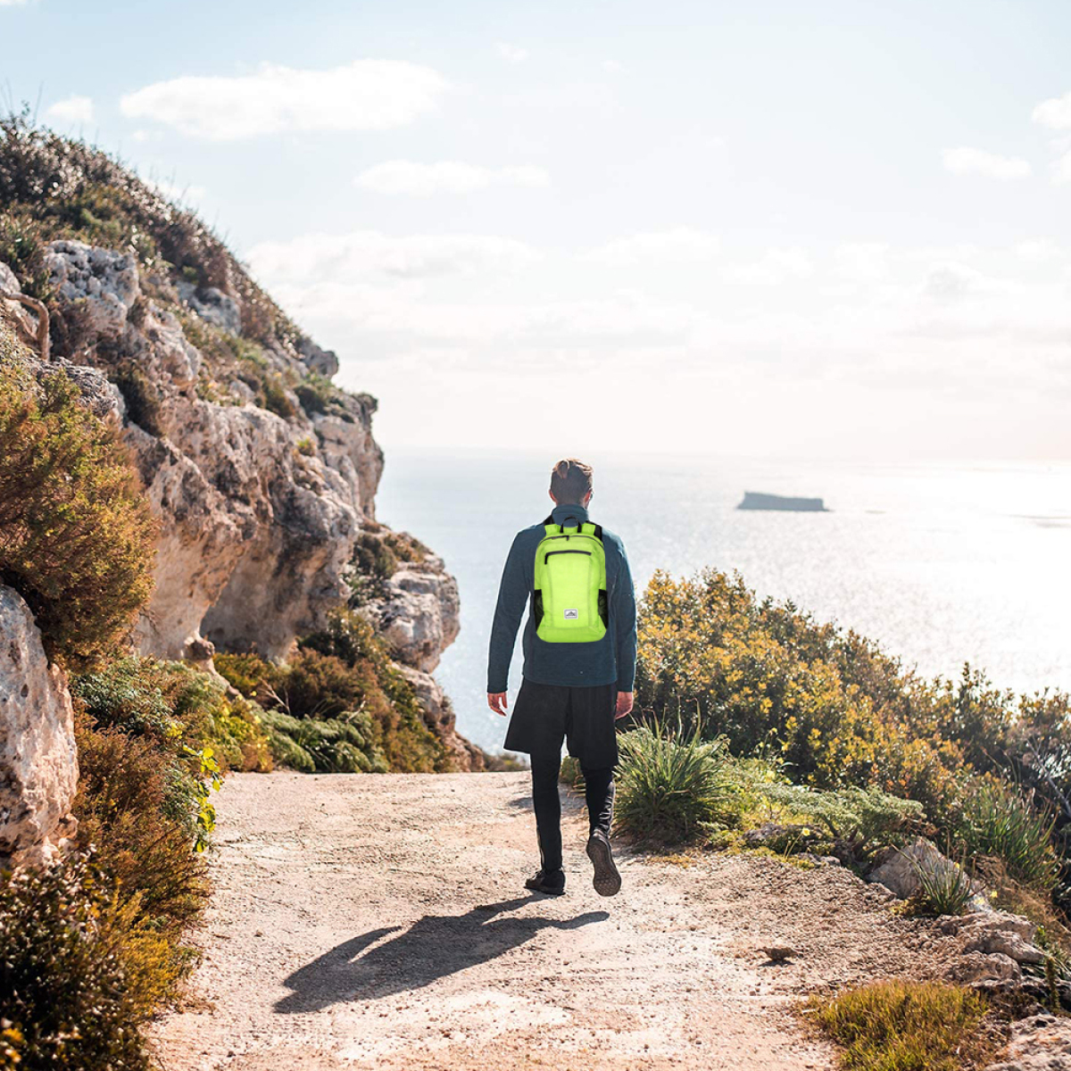 Mann geht auf einem Pfad zum Meer, mit einem grünen Rucksack. Felsiges Gelände an der Seite.