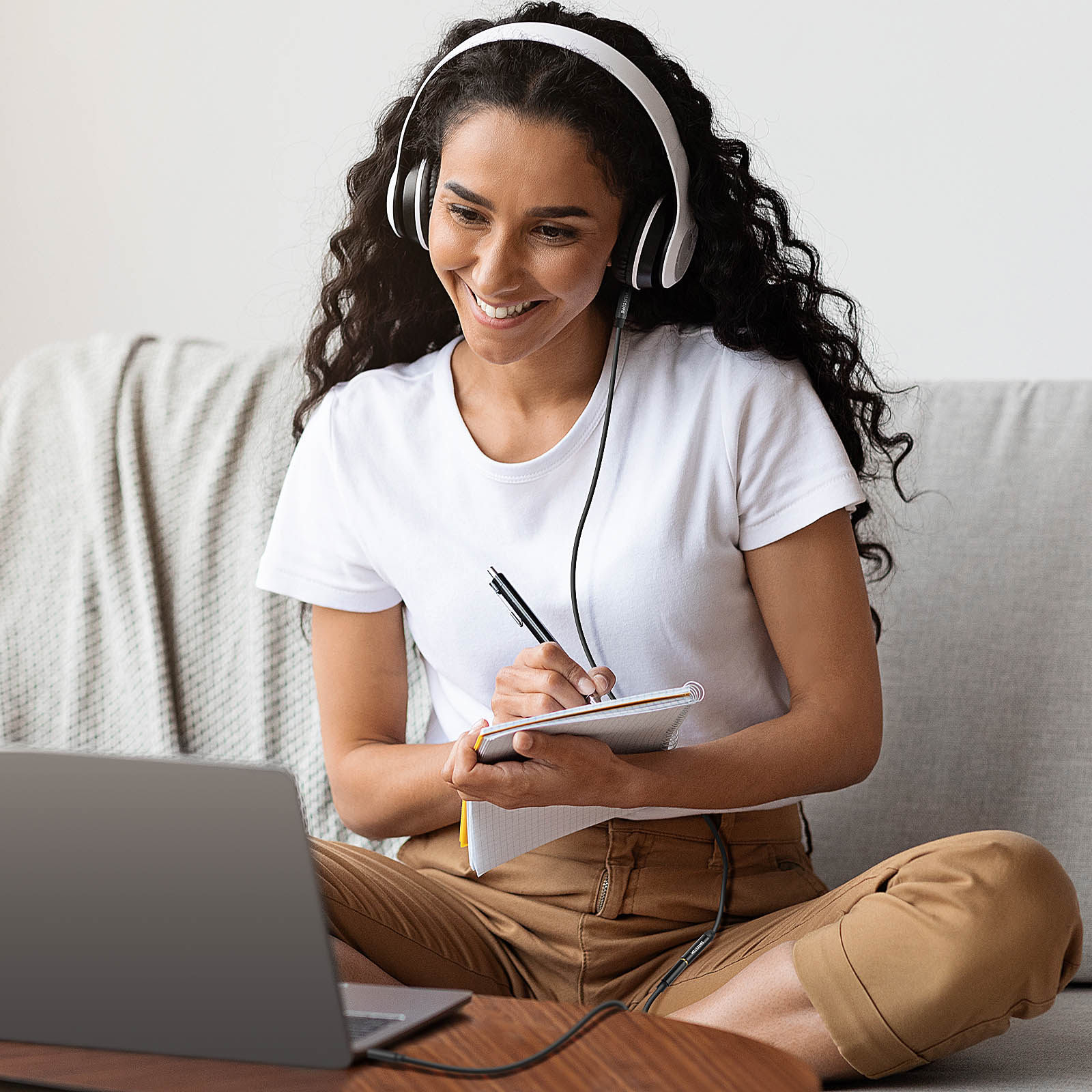 Donna con cuffie e taccuino, sorridente, guarda un laptop sul divano, ambiente interno.