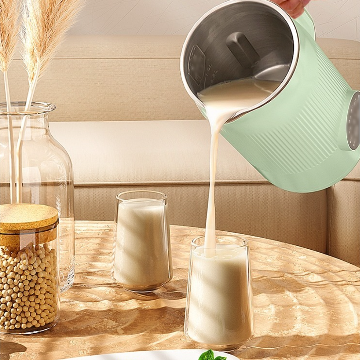 Ein grüner Sojamilchbereiter, der Milch in zwei Gläser gießt, mit Sojabohnen und einer Vase im Hintergrund.