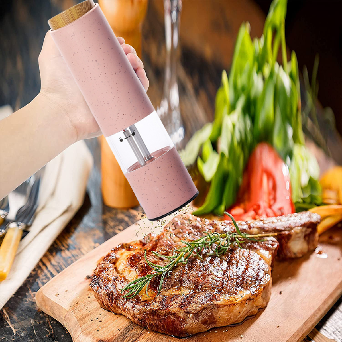 Eine Hand benutzt einen rosa elektrischen Pfeffermühle über einem Steak auf einem Holzbrett. Grüne Kräuter sind sichtbar.