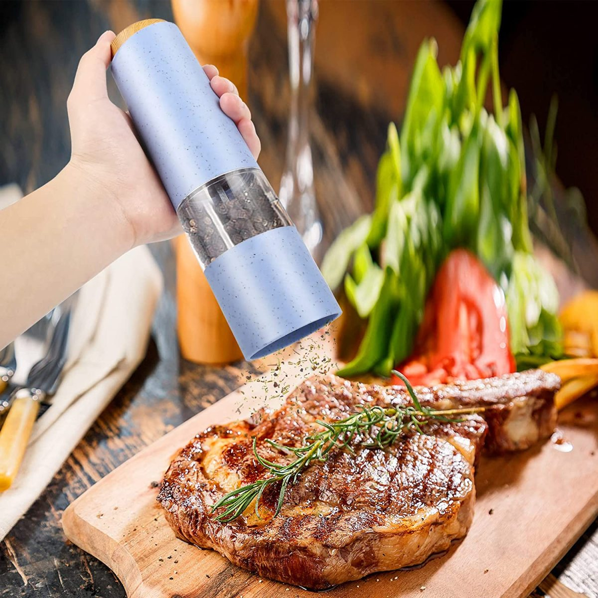 Eine Hand mahlt Pfeffer auf ein Steak auf einem Holzbrett mit Rosmarin und Grünzeug.