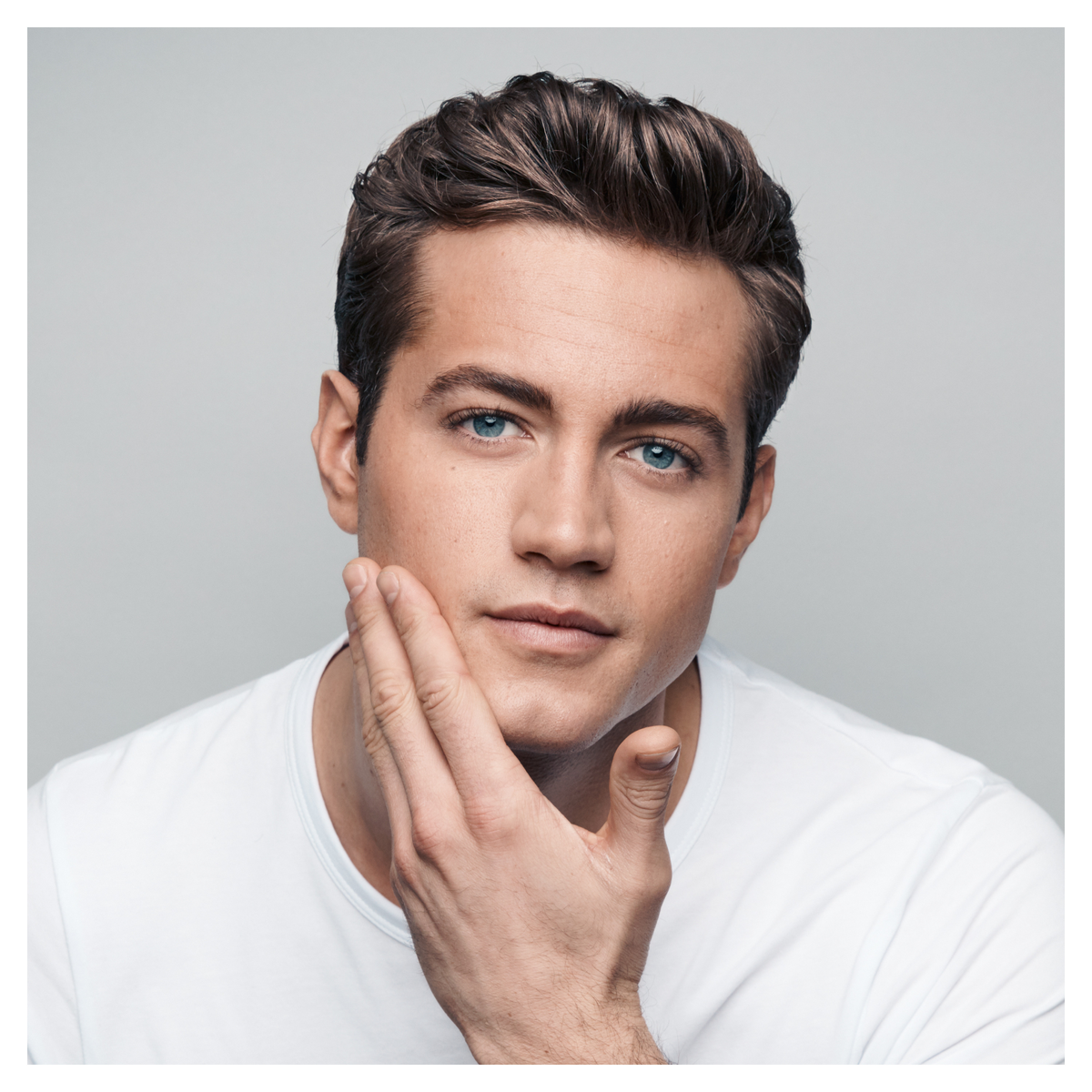 Mężczyzna z brązowymi włosami i niebieskimi oczami. Dotyka twarzy prawą ręką. Biały t-shirt.