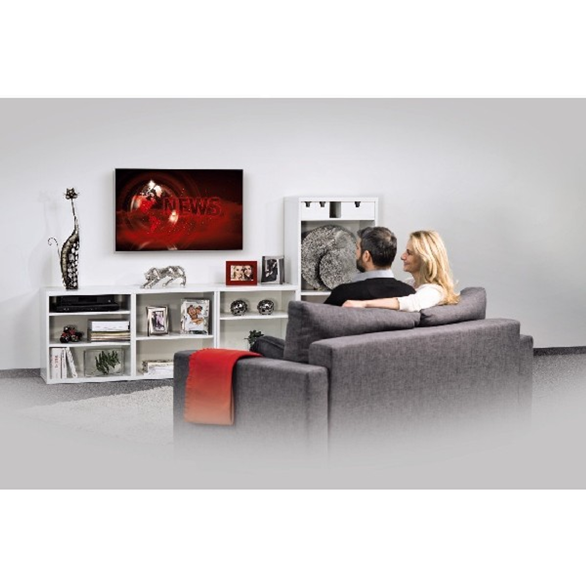 Ein Paar sieht sich eine Nachrichtensendung im Fernsehen an. Ein weißes Regal mit Dekorartikeln befindet sich auf der linken Seite.