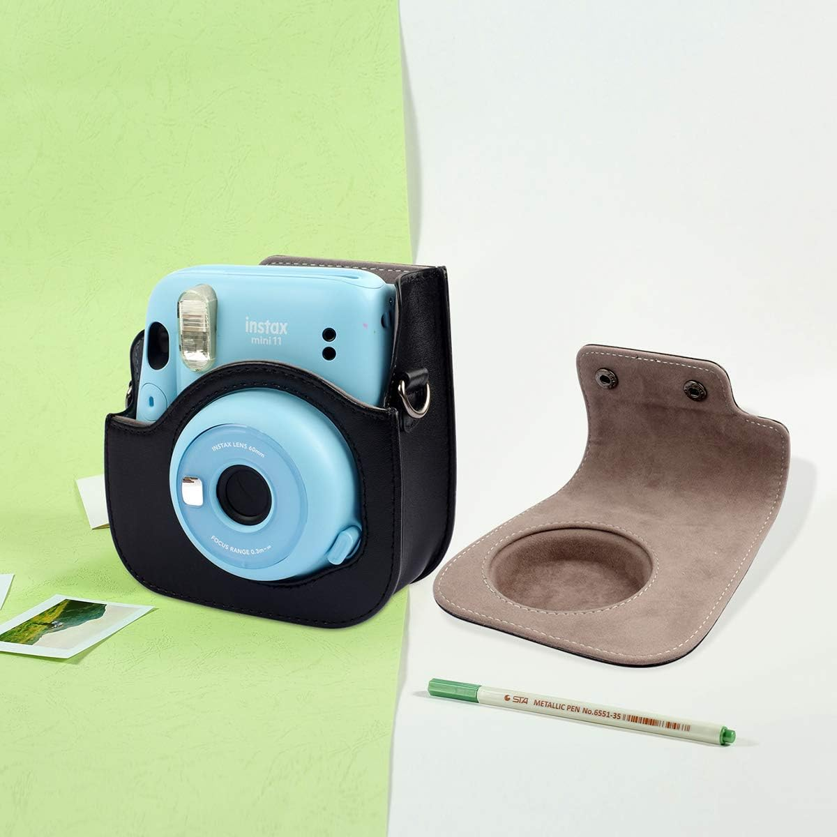 Blaue Instax Mini 11 Kamera in schwarzem Etui, offenes Etui und grüner Stift auf zweifarbigem Hintergrund.