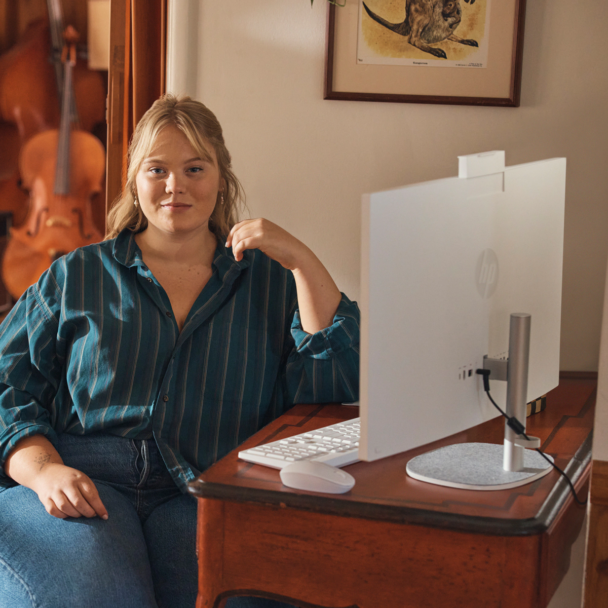 Mujer trabajando en una computadora. Sentada en un escritorio de madera, en una habitación con arte, violonchelo visible.