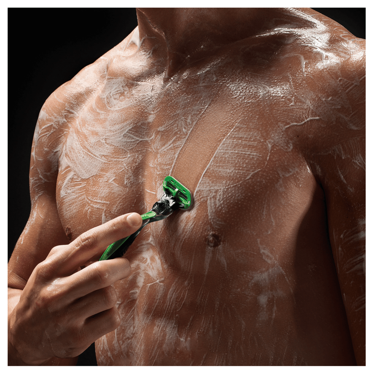 Hombre afeitándose el pecho con una maquinilla verde. Crema de afeitar blanca visible. Fondo negro.