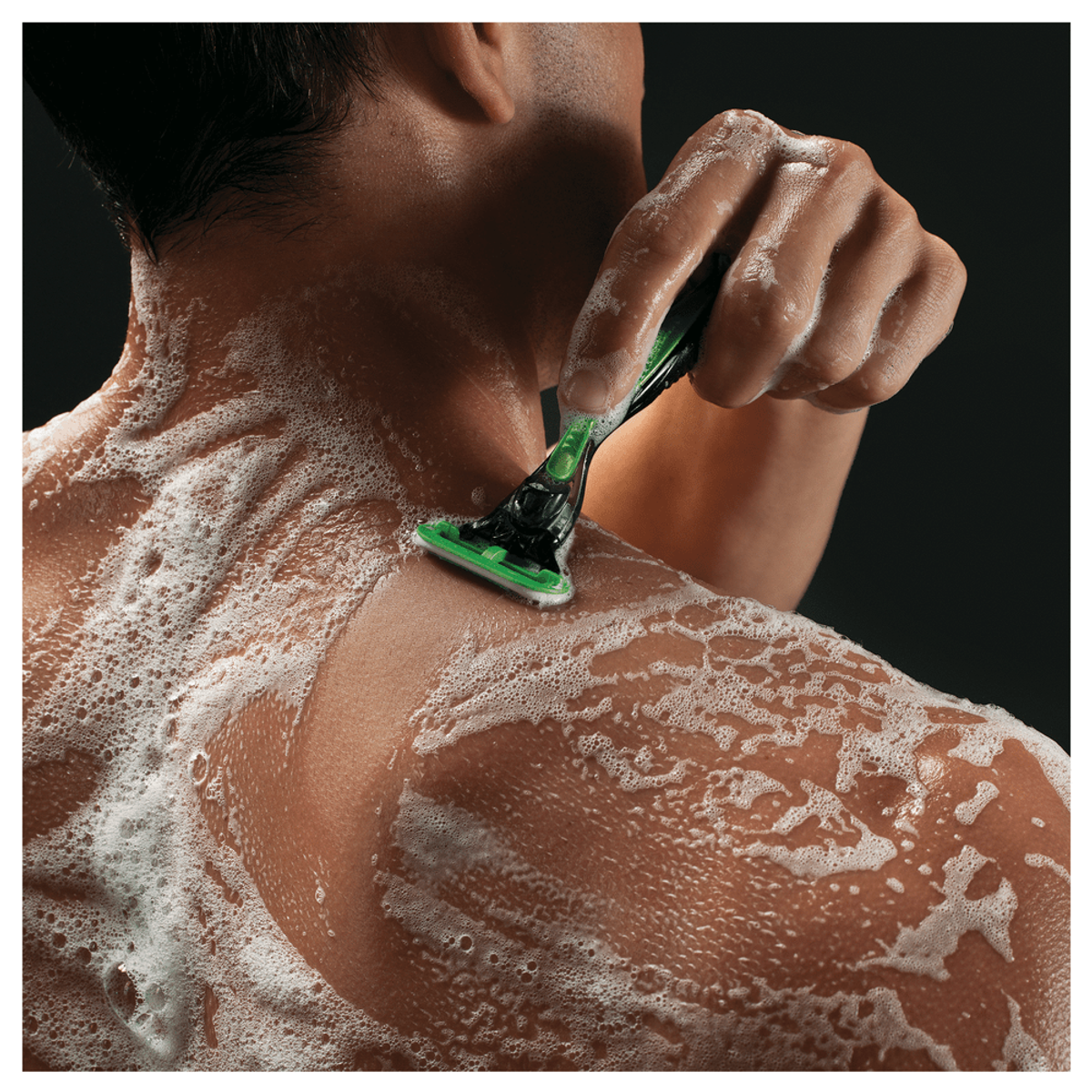 Hombre afeitándose la espalda con una maquinilla verde. La espuma cubre su piel y la maquinilla.