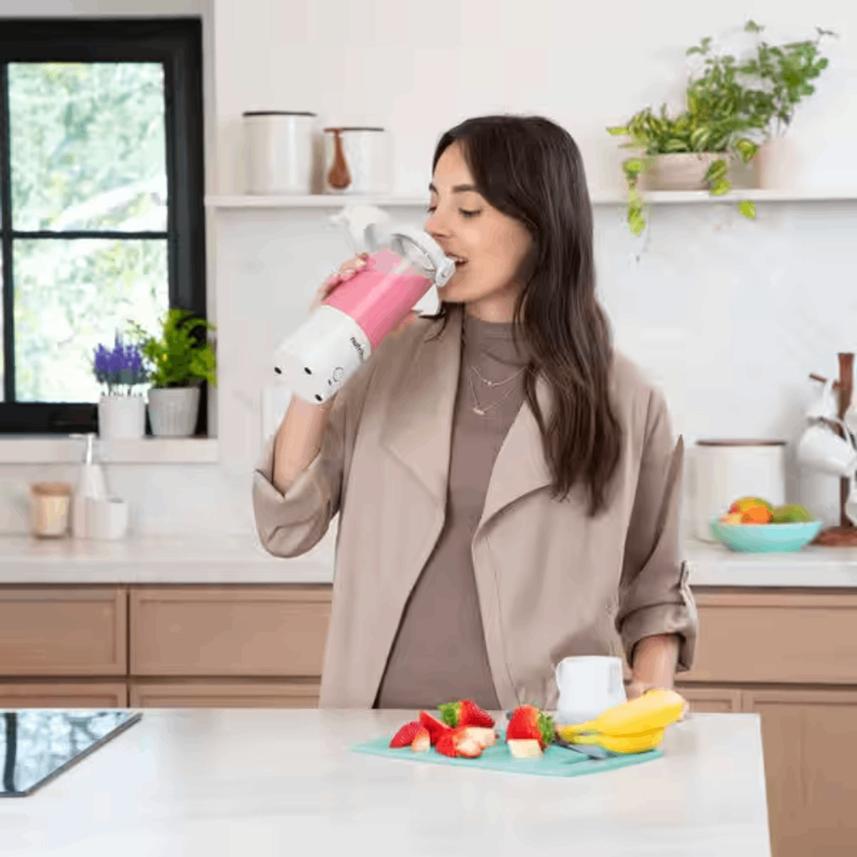 Kobieta pije z różowego blendera. Truskawki i banan na blacie. Otoczenie kuchni, naturalne światło.