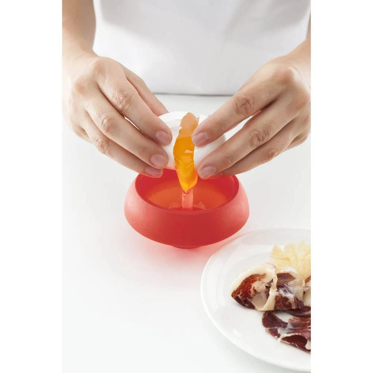 Mani che rompono un uovo in un separatore rosso con un piatto di carne e formaggio.