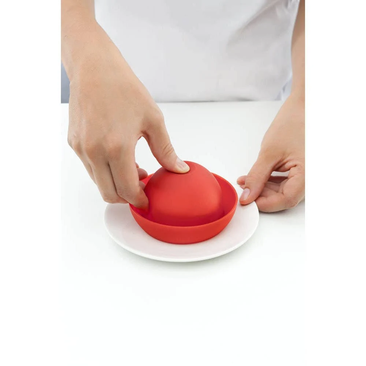 Mani che posizionano una ciotola di silicone rossa all'interno di un piatto bianco. Sfondo bianco.