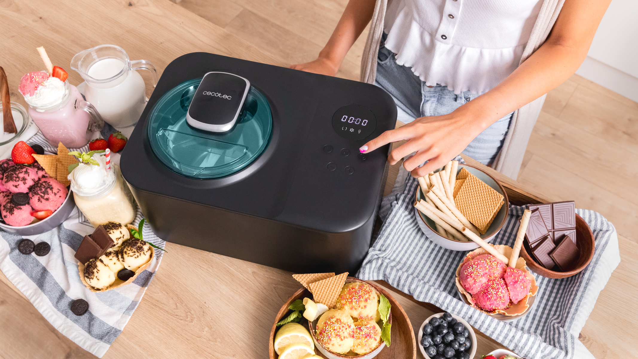 Kobieta obsługuje czarną maszynę do lodów, otoczona miseczkami z lodami, dodatkami i słodyczami.