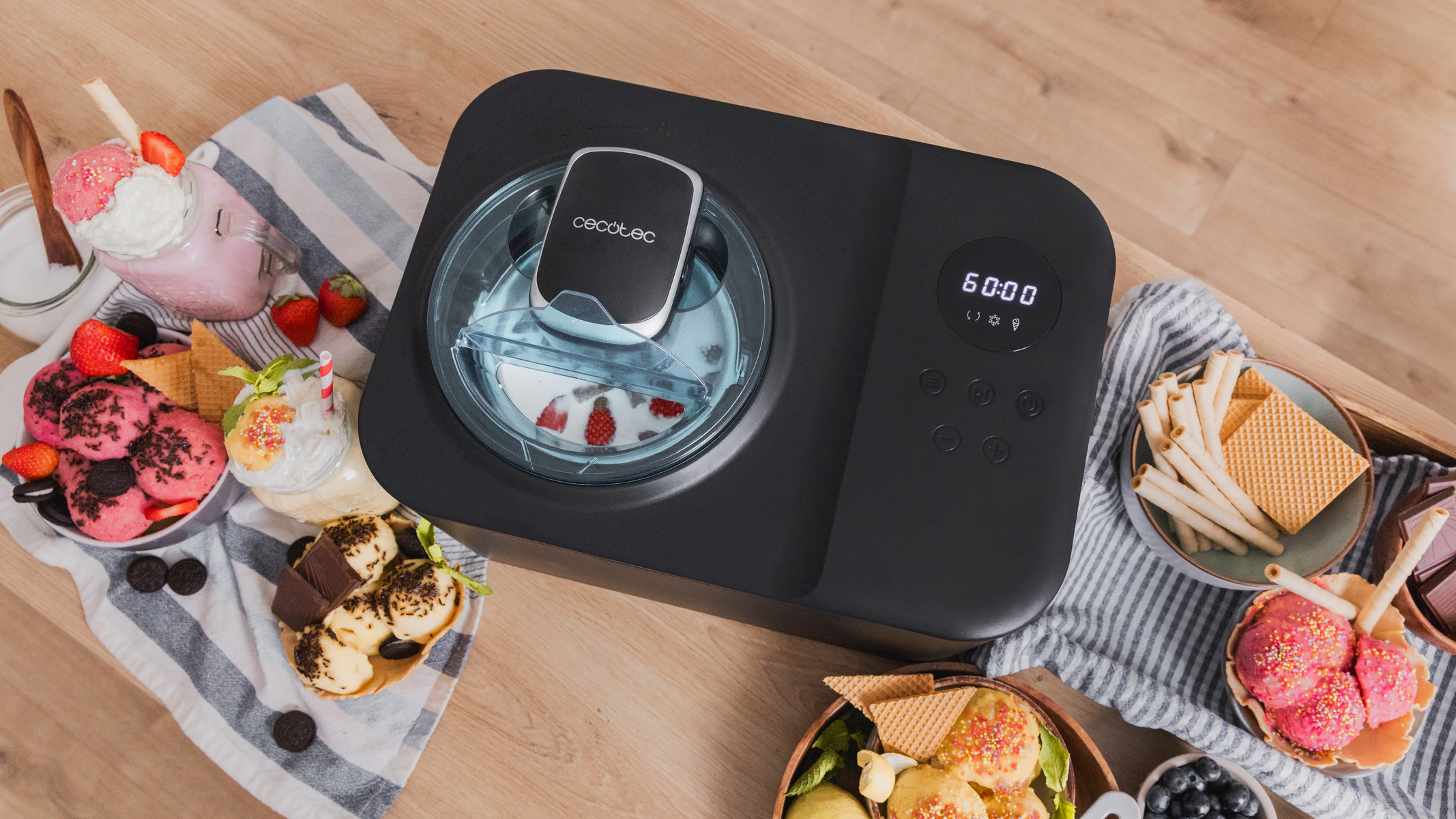 Maszyna do lodów z deserami wokół. Zawiera lody, gofry, truskawki i bitą śmietanę. Na drewnianym stole.