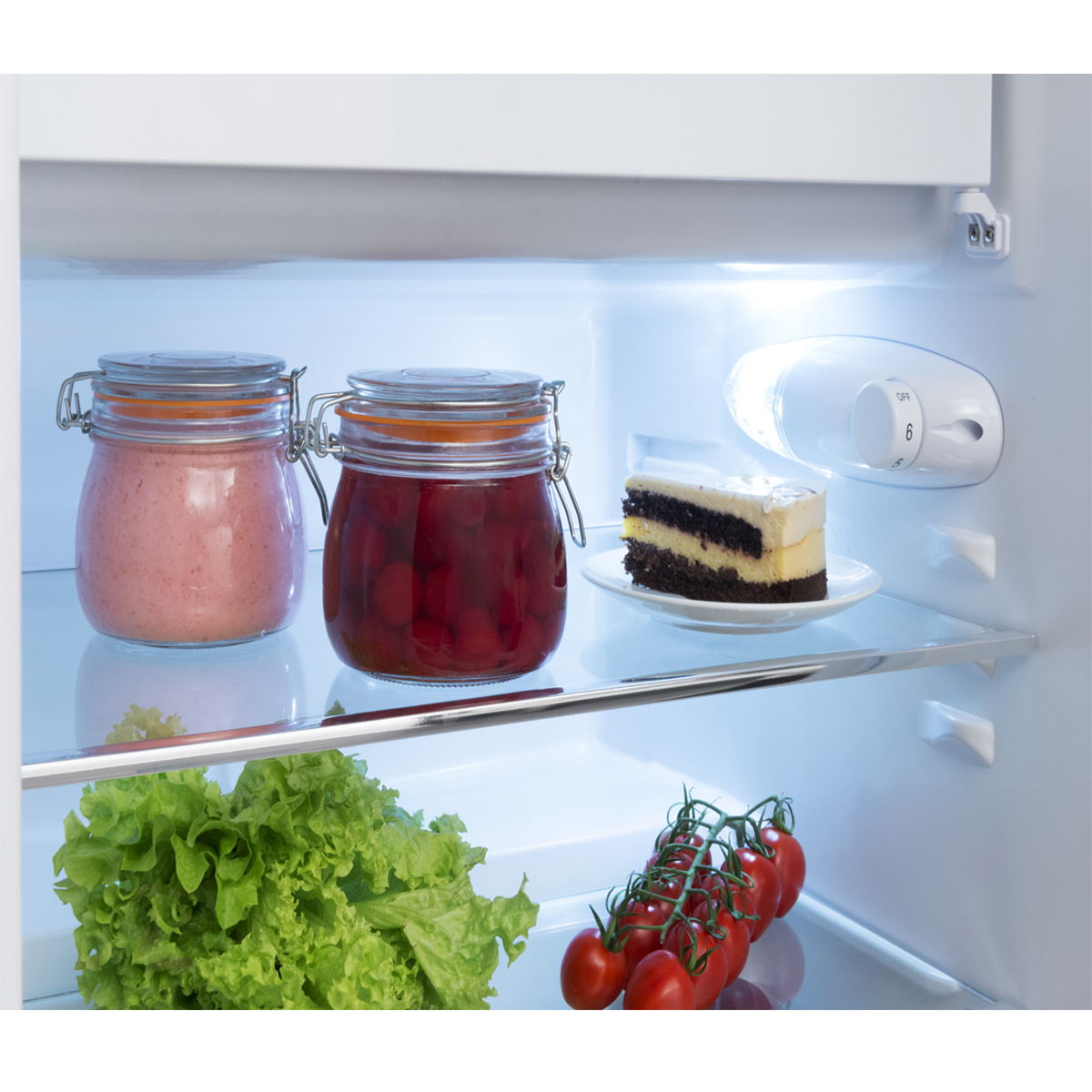 In einem Kühlschrank: Gläser mit rosa und roten Konserven, Kuchen auf einem Teller, Salat, Tomaten.