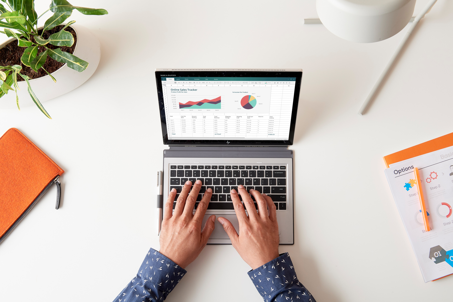Osoba korzystająca z tabletu z klawiaturą na białym biurku. Arkusz kalkulacyjny na ekranie. Widoczna roślina i notatnik.
