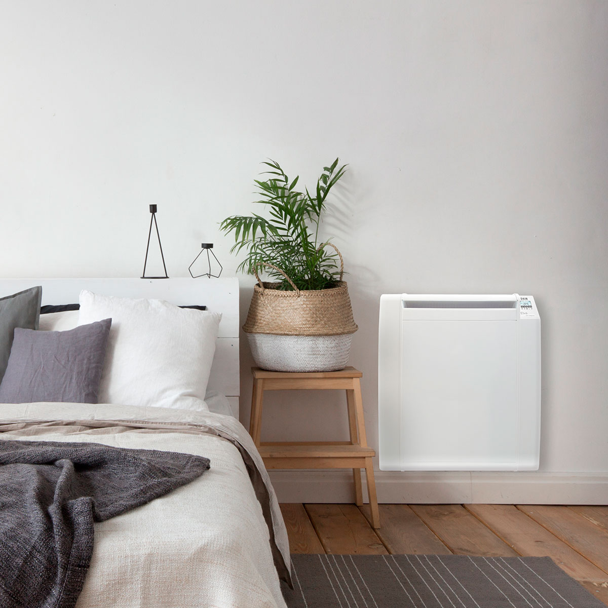 Calentador blanco montado en la pared junto a una cama. Planta en cesta sobre taburete.