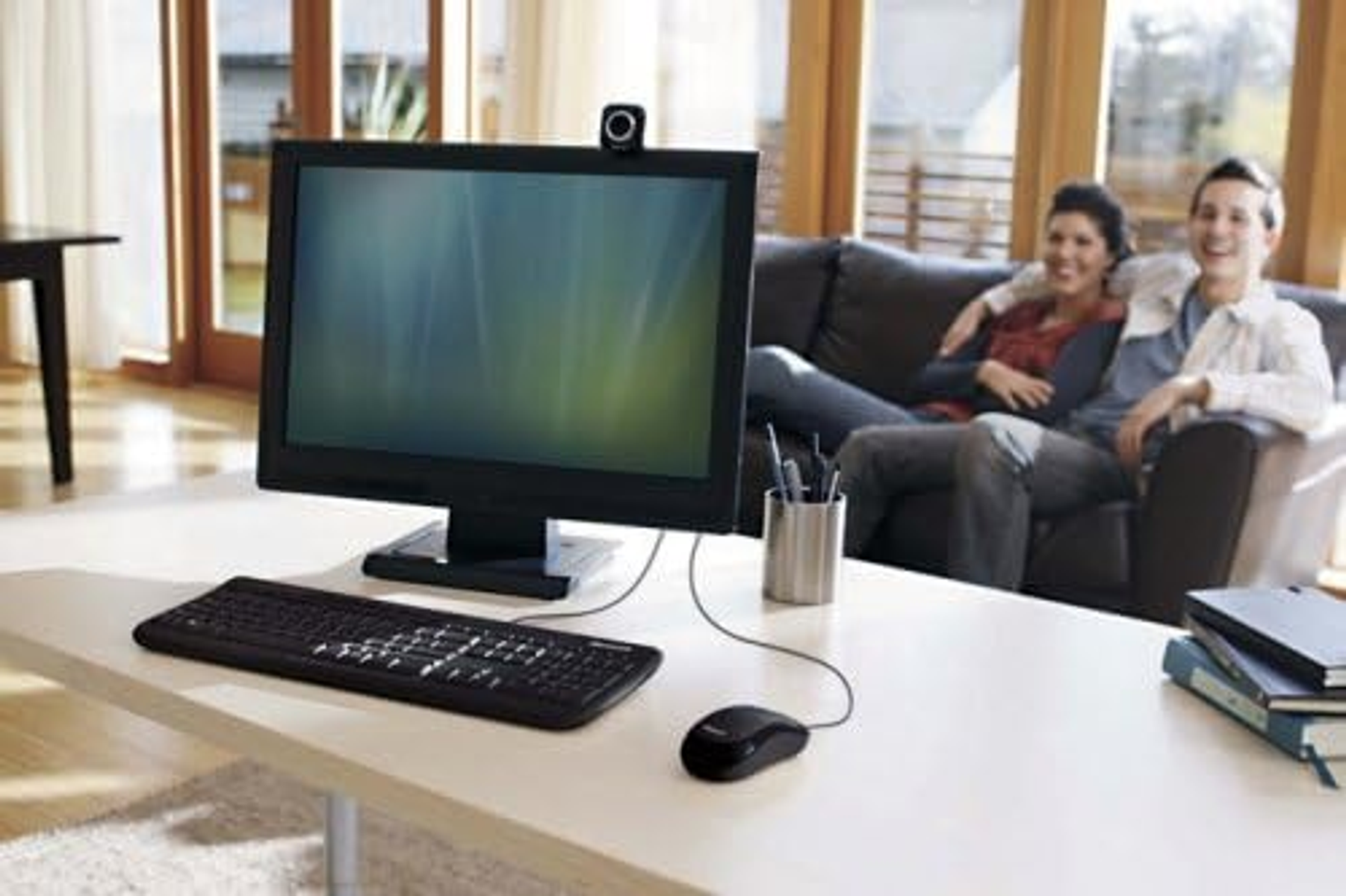 Ein Computer-Setup auf einem Tisch, mit Monitor, Tastatur, Maus und zwei Personen im Hintergrund.