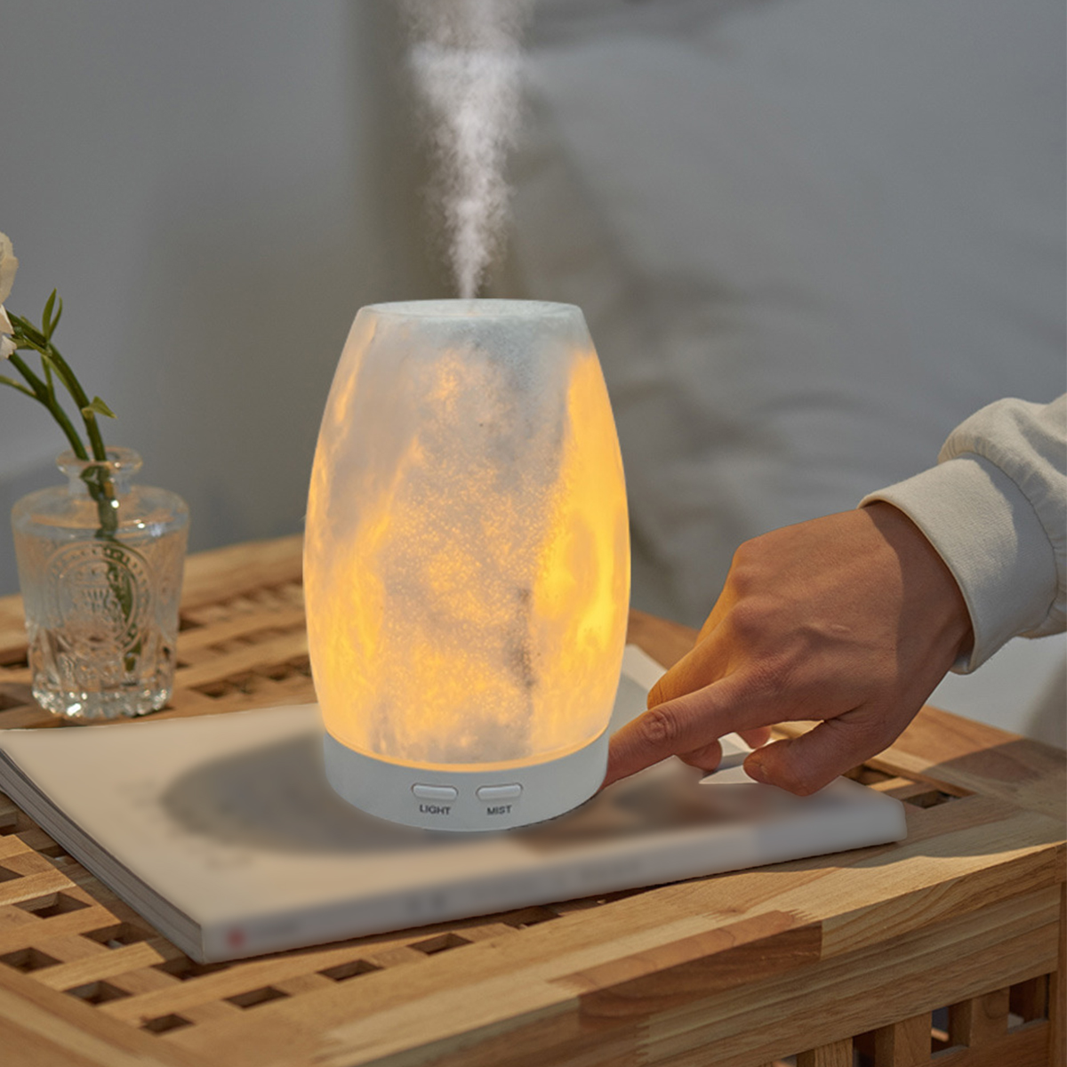 Ein Aromadiffusor mit einer Hand, die einen Knopf drückt. Er steht auf einem Buch auf einem Holztisch.