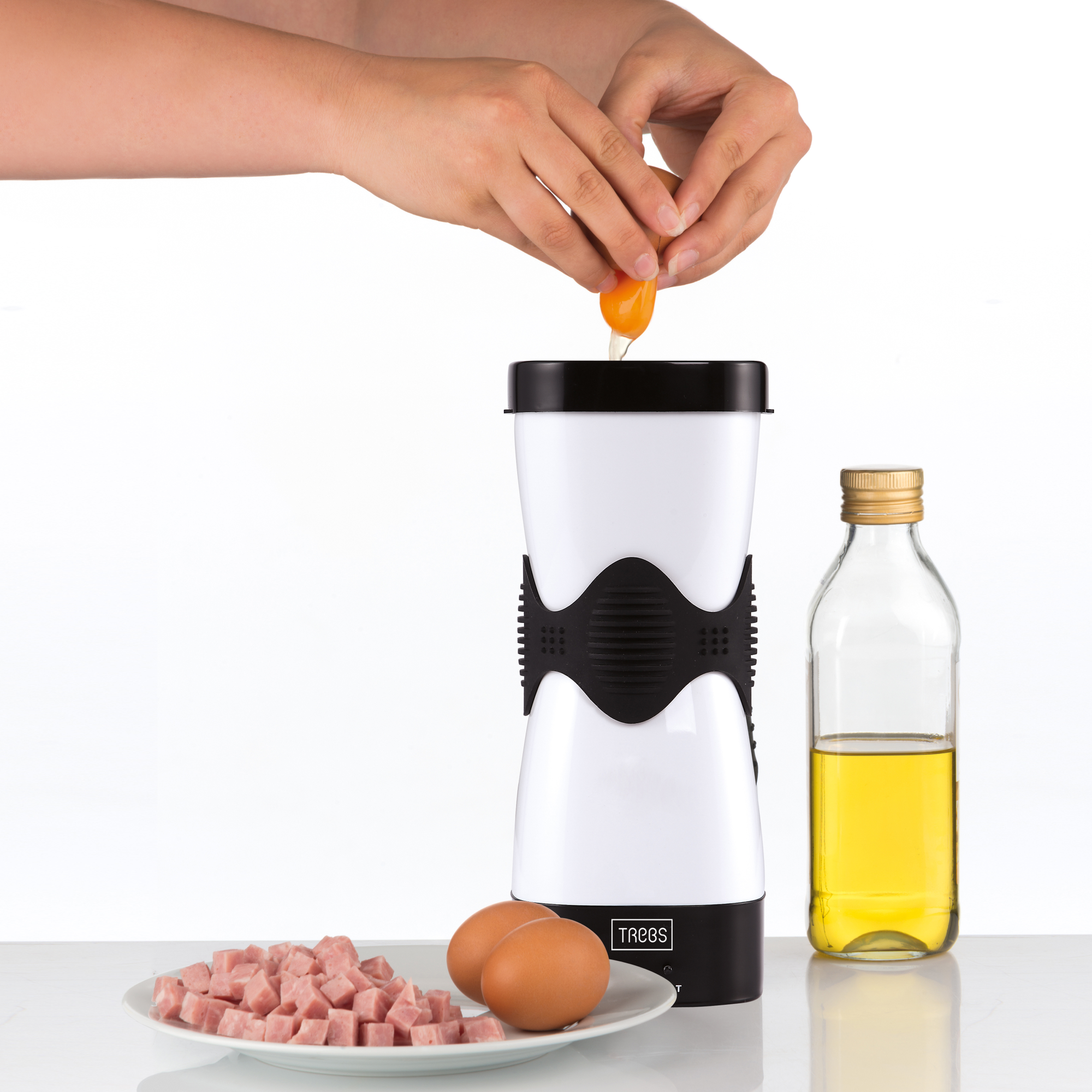 Mani che rompono un uovo in un'apparecchiatura per uova. Accanto ci sono uova, olio d'oliva e cubetti di prosciutto su un piatto.