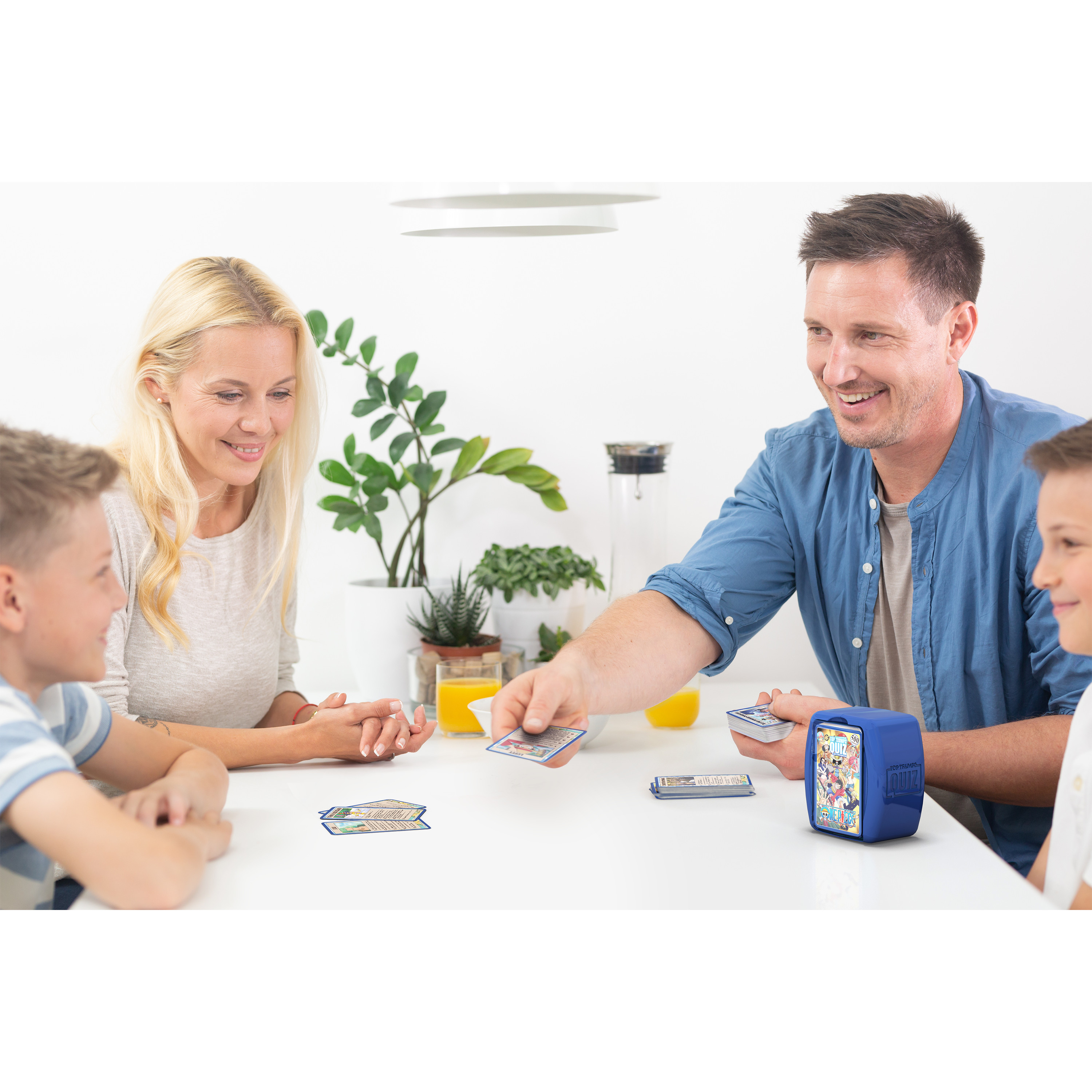 Familie spielt ein Kartenspiel. Der Tisch hat eine blaue Box. Die Szene ist in einem hellen Raum.