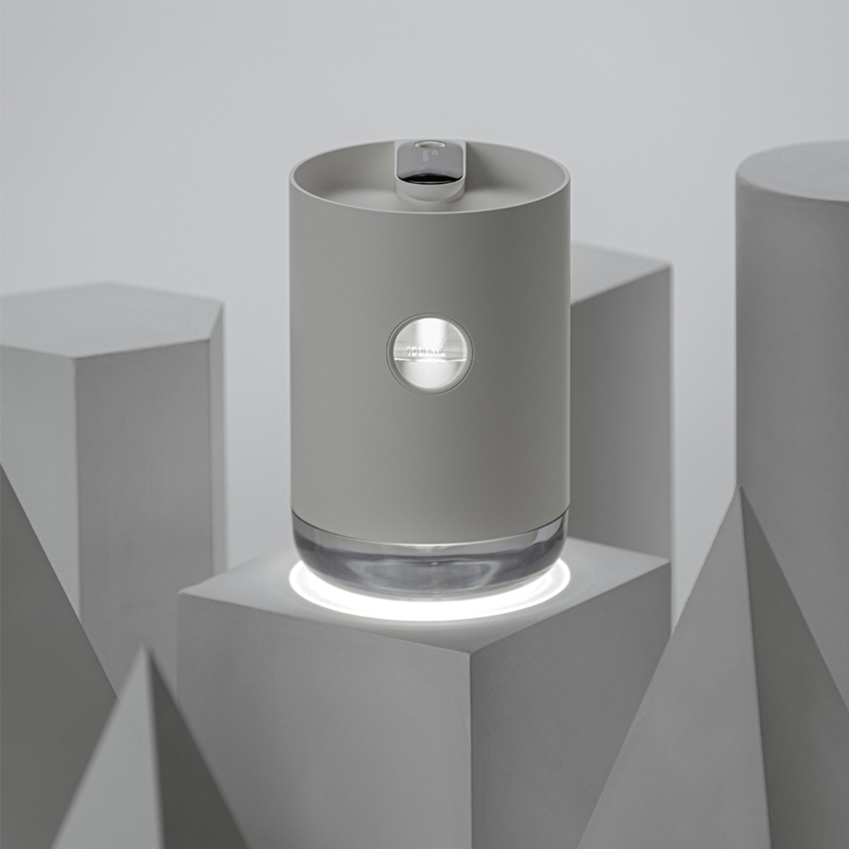 Ein graues, zylindrisches Gerät mit hellem Sockel, platziert auf geometrischen weißen Formen.