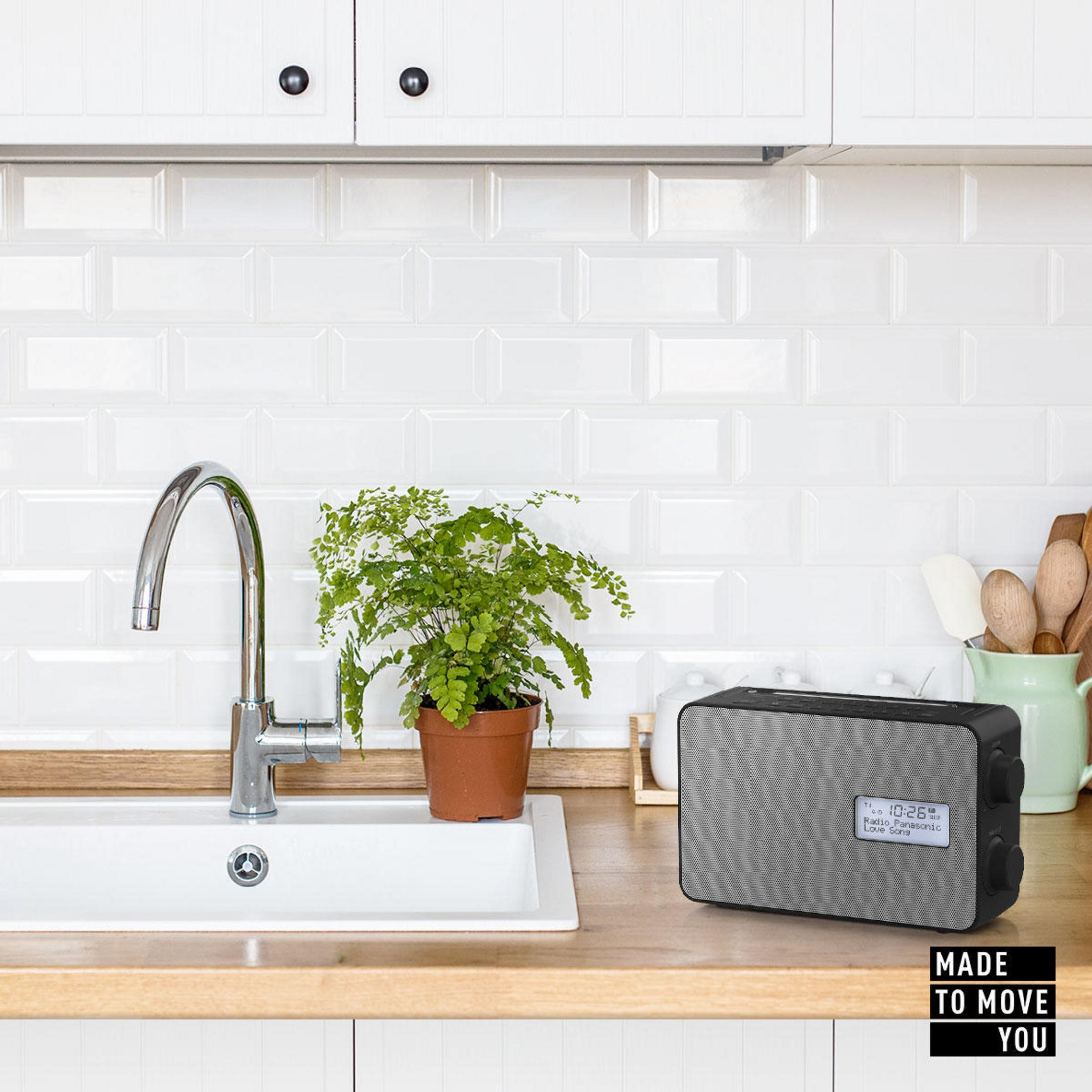 Radio na drewnianym blacie, ze zlewem, rośliną i przedmiotami kuchennymi w tle.