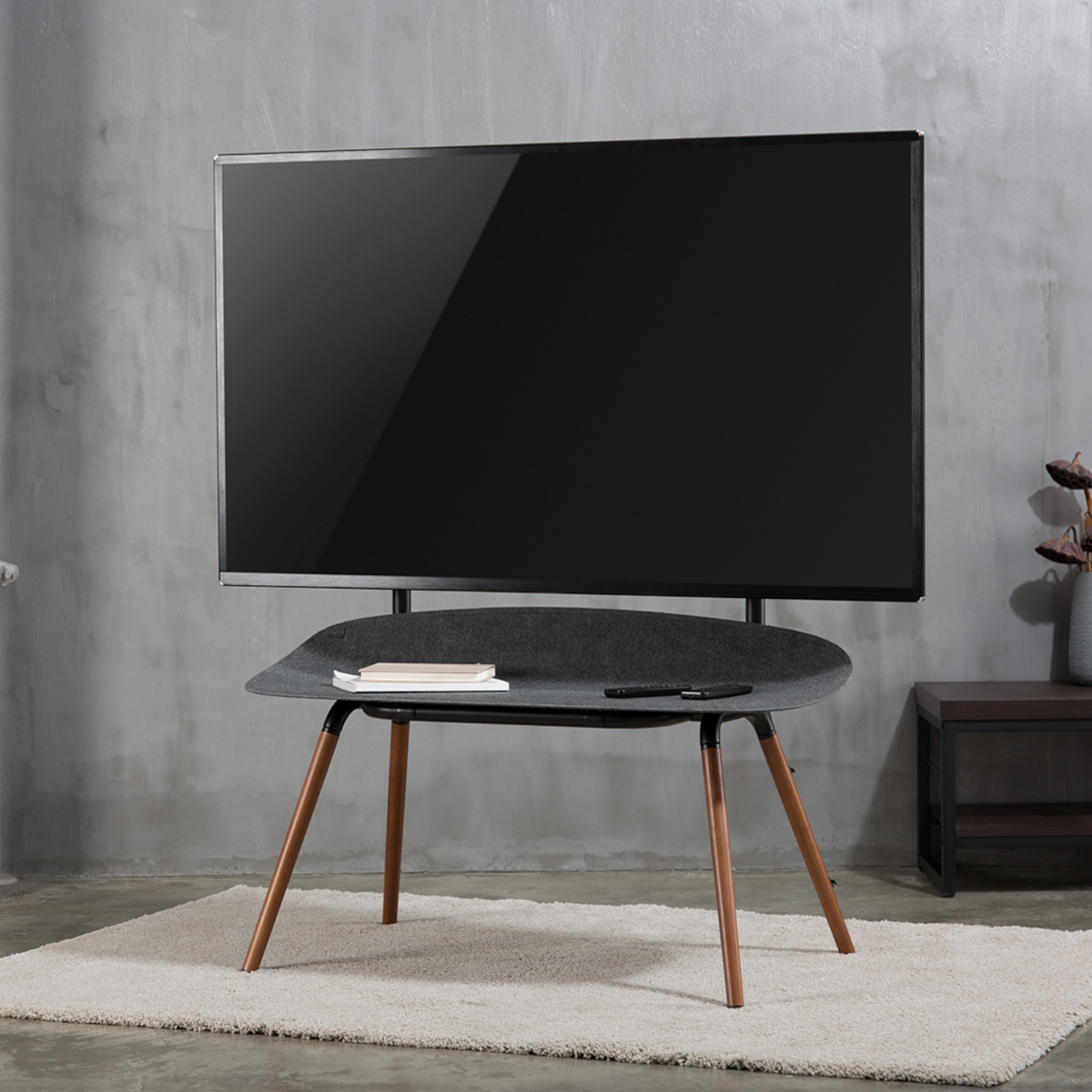 Ein schwarzer Fernseher auf einem Ständer mit Holzbeinen, mit einem Teppich davor und einem grauen Hintergrund.