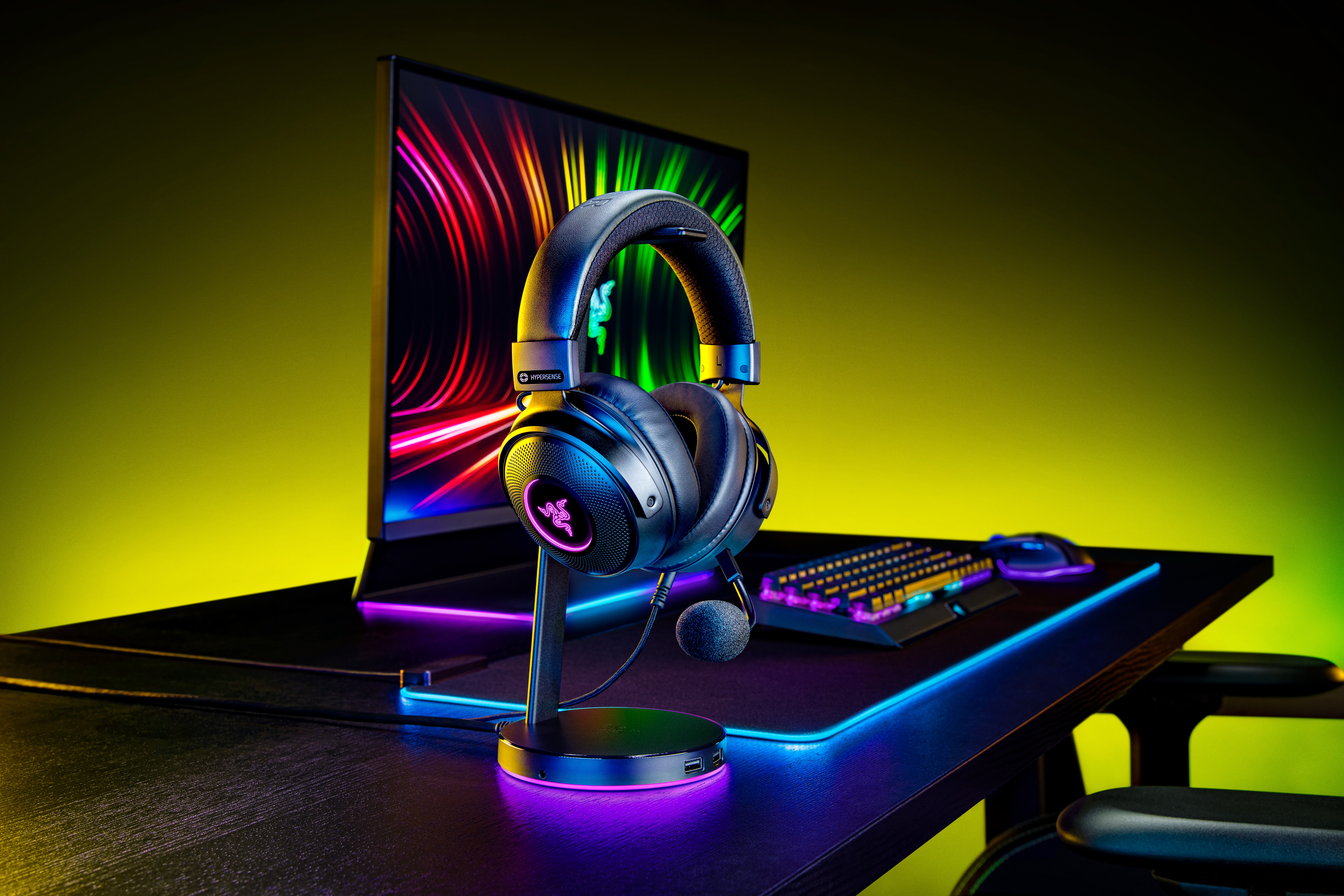 Schwarzes Gaming-Headset auf Ständer. Hintergrund: Monitor, Tastatur, Maus, bunte Lichter.