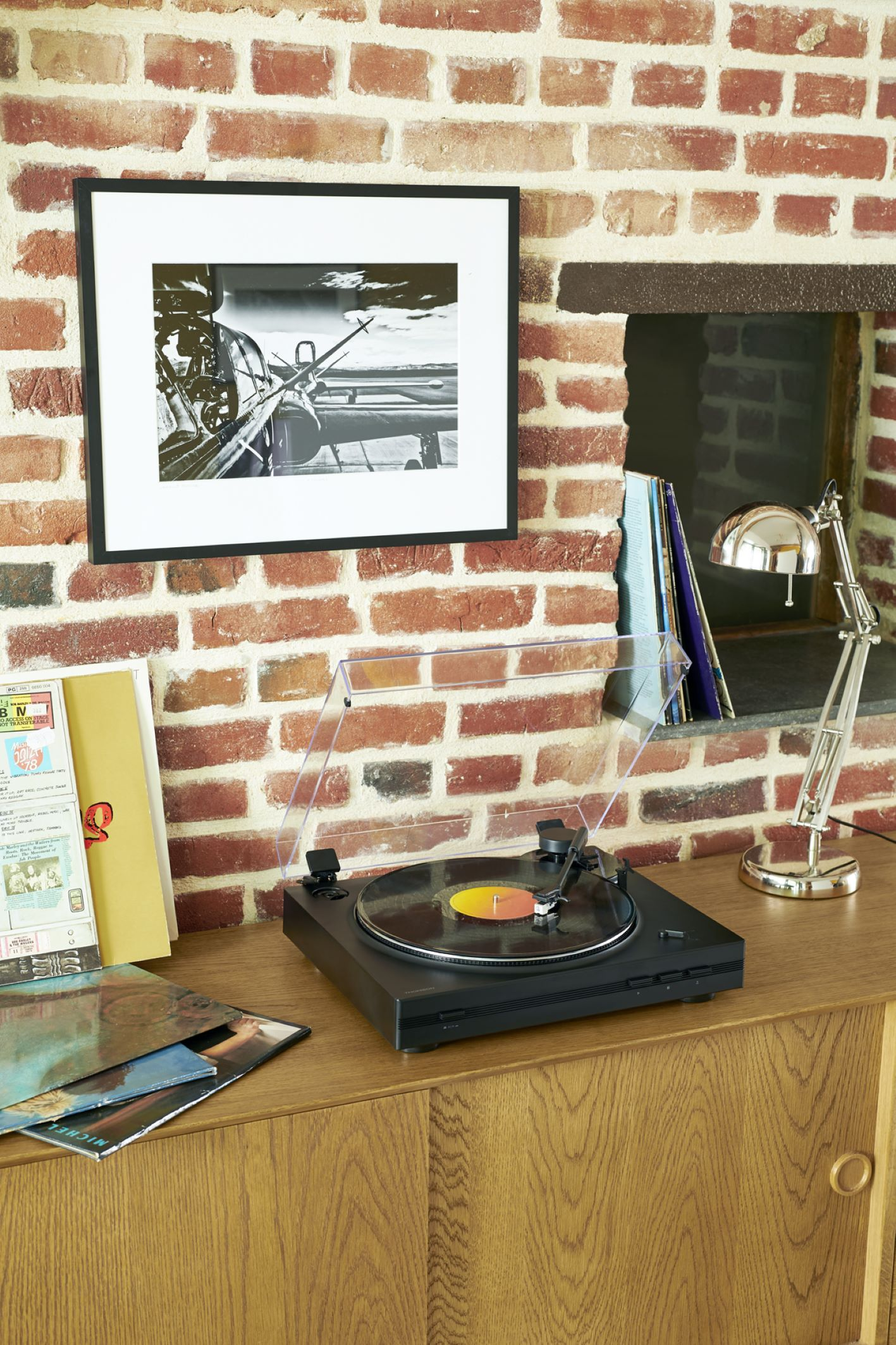 Un tocadiscos en un mueble de madera. Una imagen con marco negro y una lámpara están en el fondo.