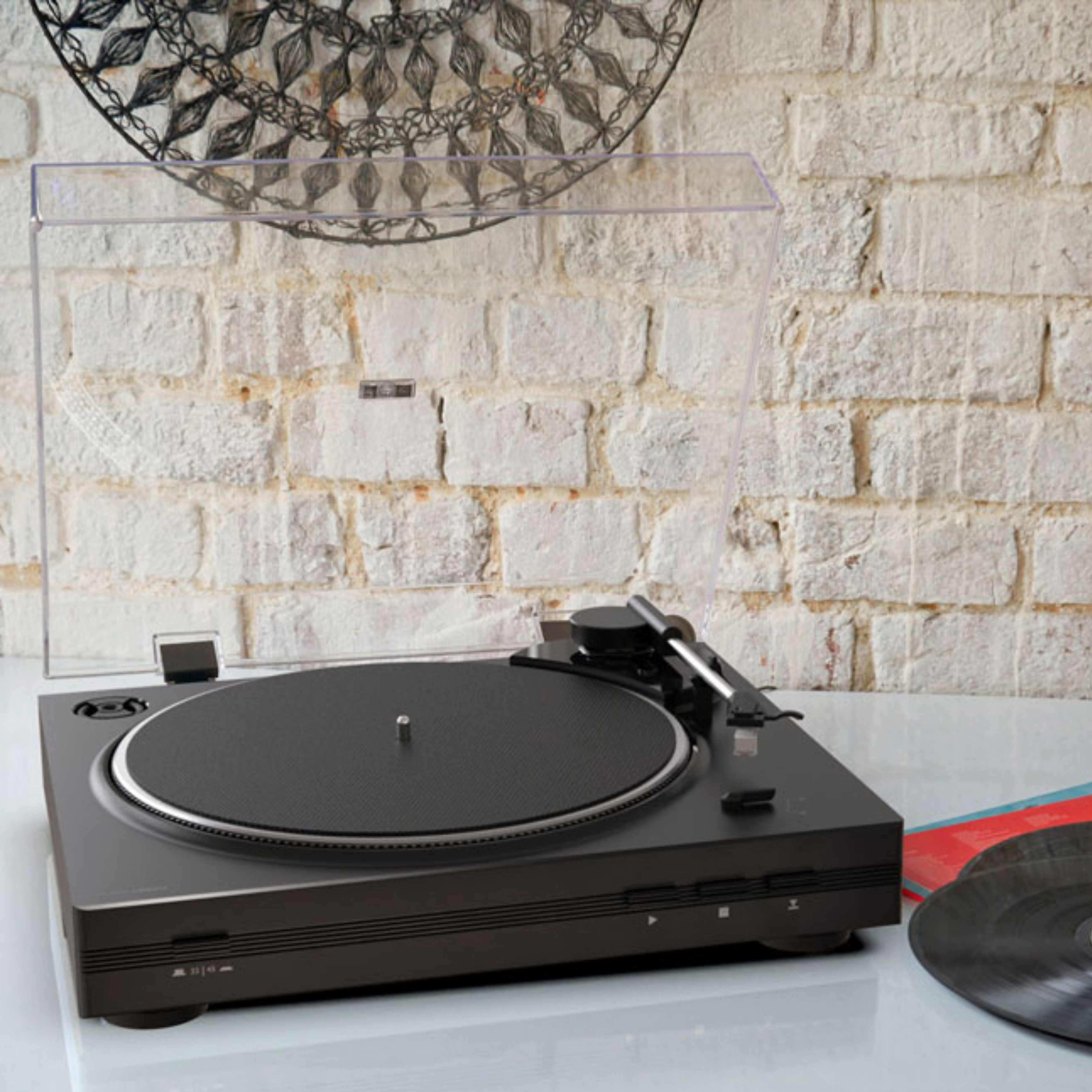 Gramofon na białym stole, tło z ceglanej ściany. Czarne płyty winylowe po prawej.