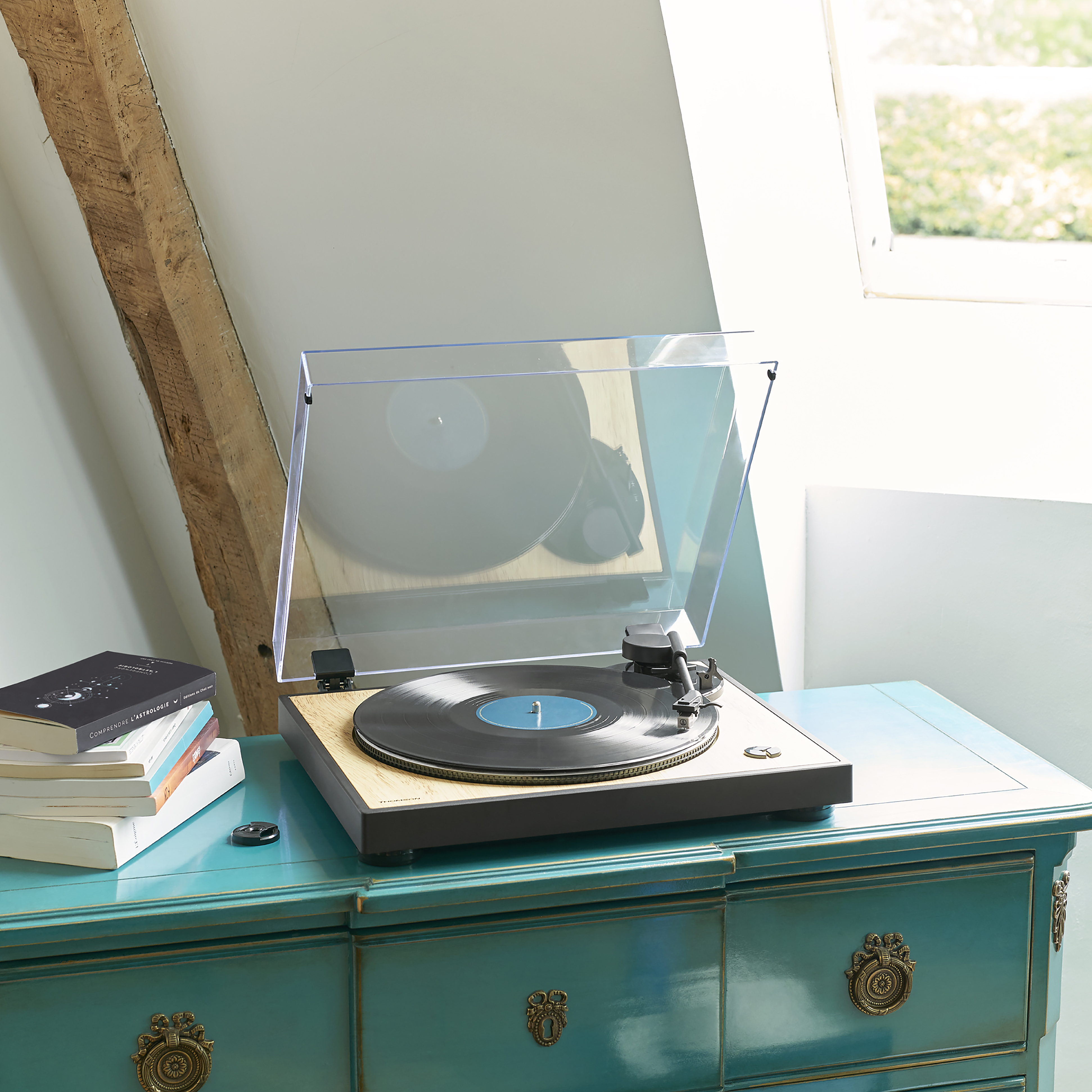 Gramofon na niebieskiej szafce, ze stosem książek i oknem w tle.