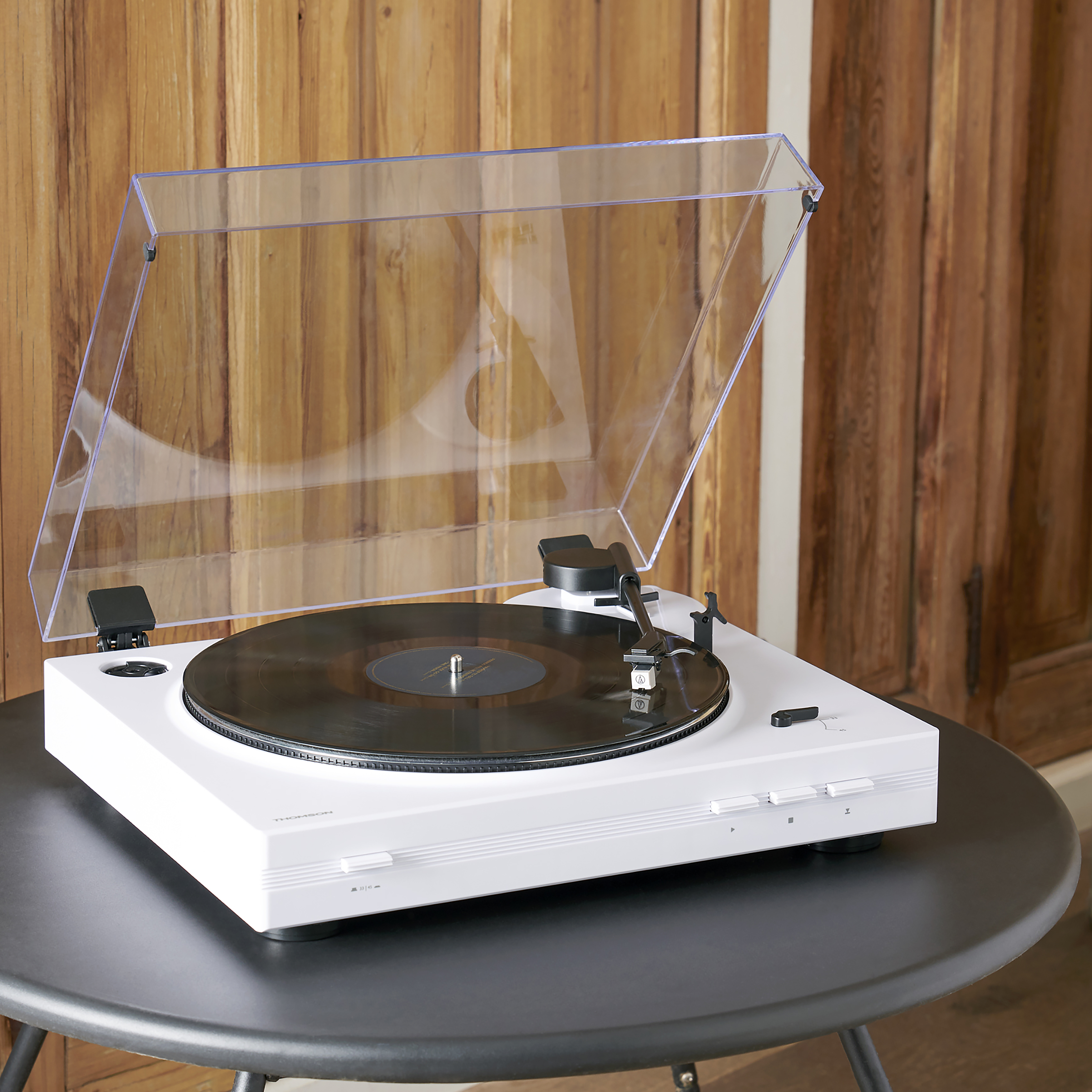 Biały gramofon z przezroczystą pokrywą, na czarnym stole, obok drewnianej ściany.