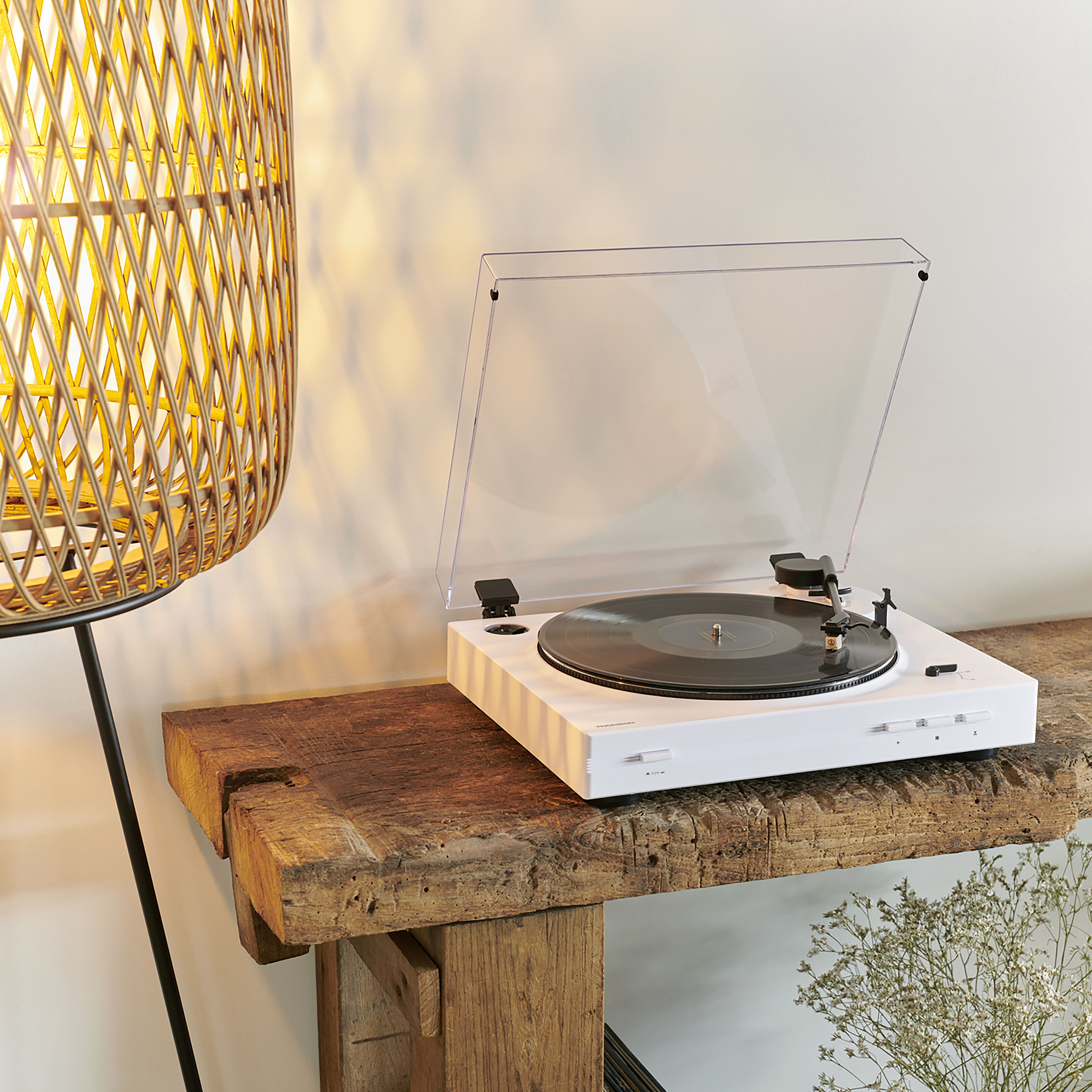 Biały gramofon na drewnianym stole, z otwartą przezroczystą pokrywą. W pobliżu tkana lampa i kwiaty.