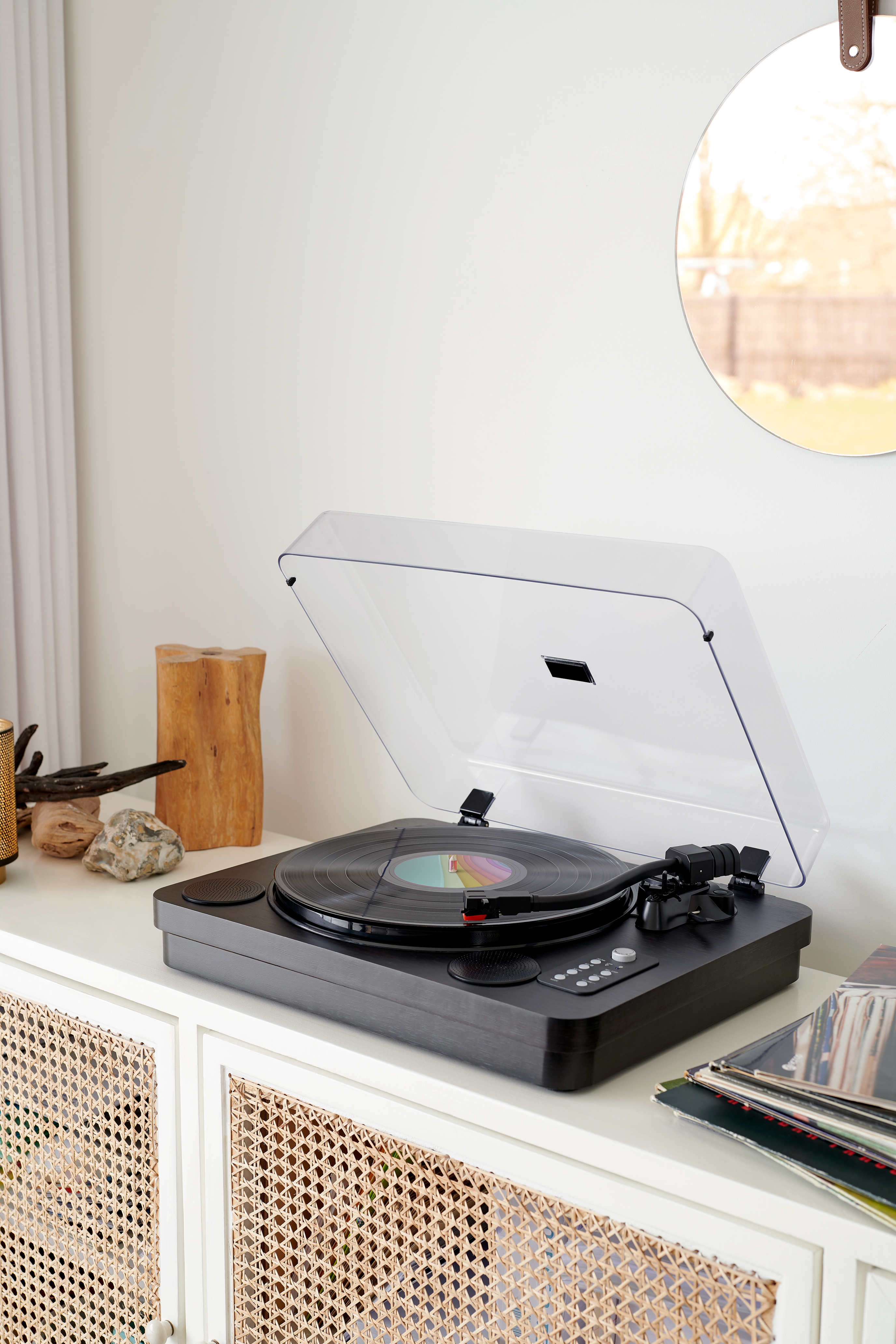 Czarny gramofon z otwartą przezroczystą pokrywą, na białej szafce, w pobliżu lustra i albumów.