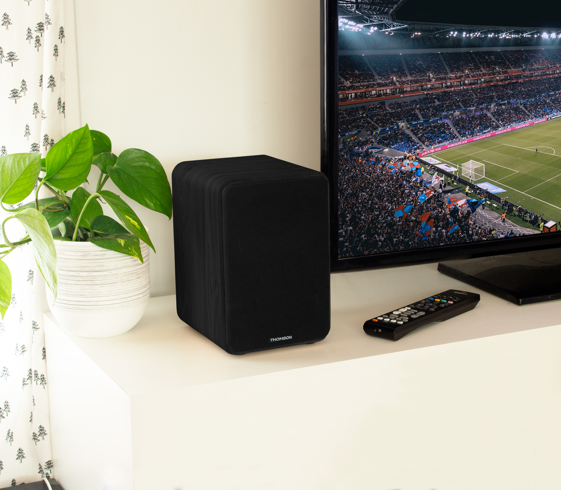 Czarny głośnik na białej powierzchni. Telewizor po prawej stronie z meczem piłki nożnej. Roślina doniczkowa po lewej.