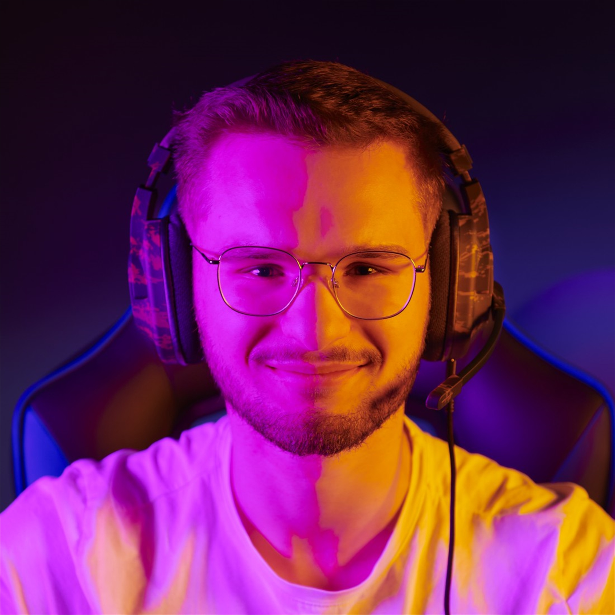 Ein lächelnder Mann mit Brille und Kopfhörern, beleuchtet von rosa und gelben Lichtern, ist im Bild.