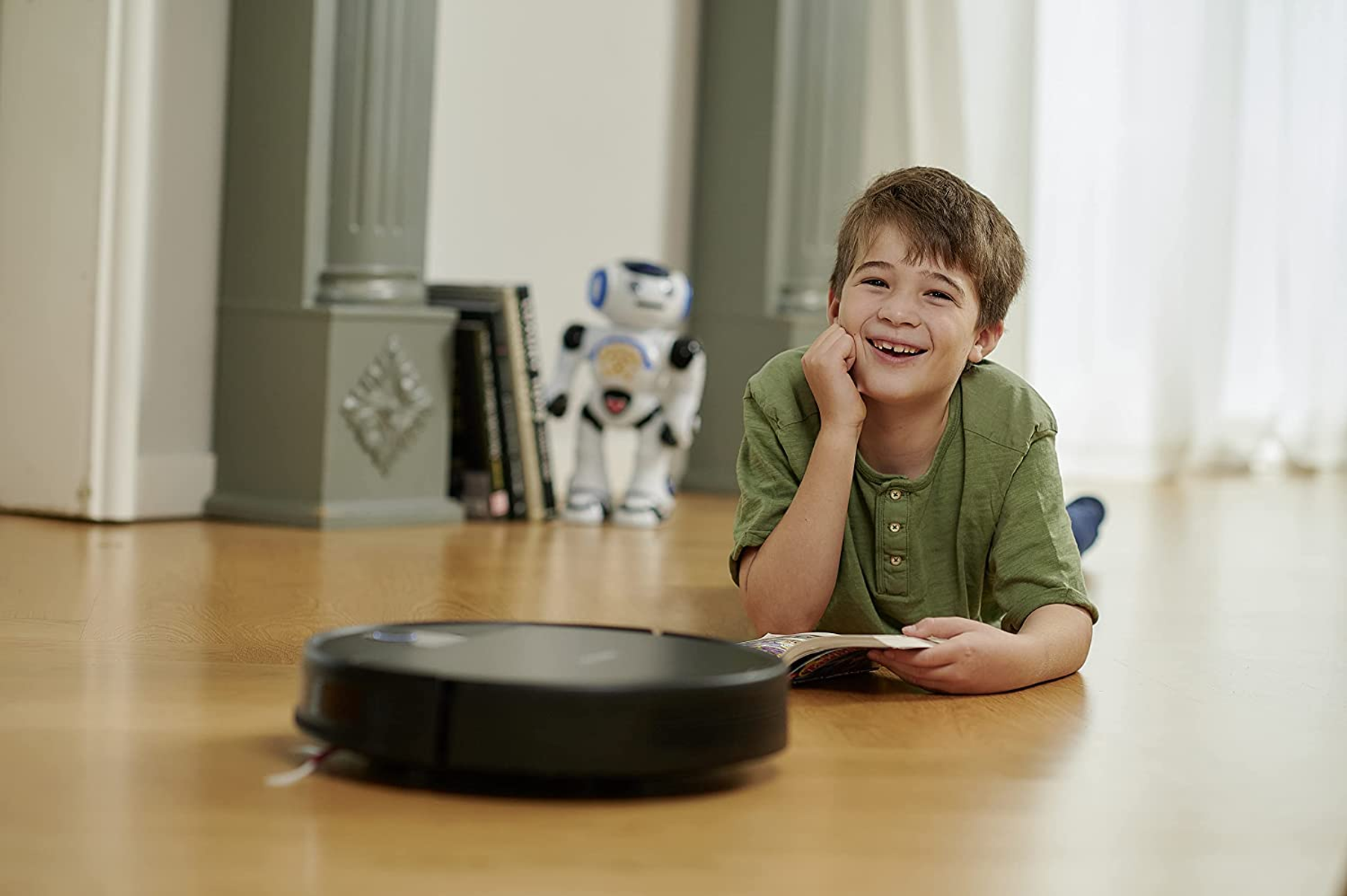 Ragazzo sdraiato sul pavimento, con un libro, sorridente. Robot e aspirapolvere sullo sfondo.
