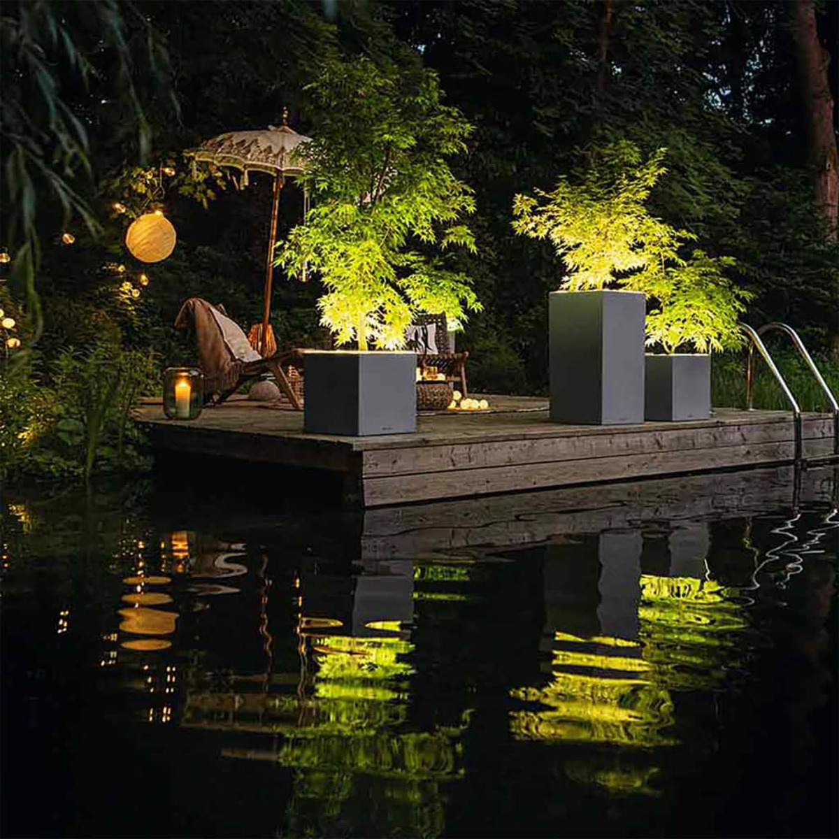 Un ponton en bois avec de l'eau sombre et des éléments paysagers illuminés la nuit.