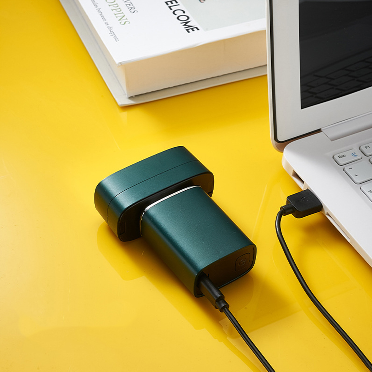 Grüne Powerbank, verbunden mit einem weißen Laptop auf gelber Oberfläche. USB-Kabel.