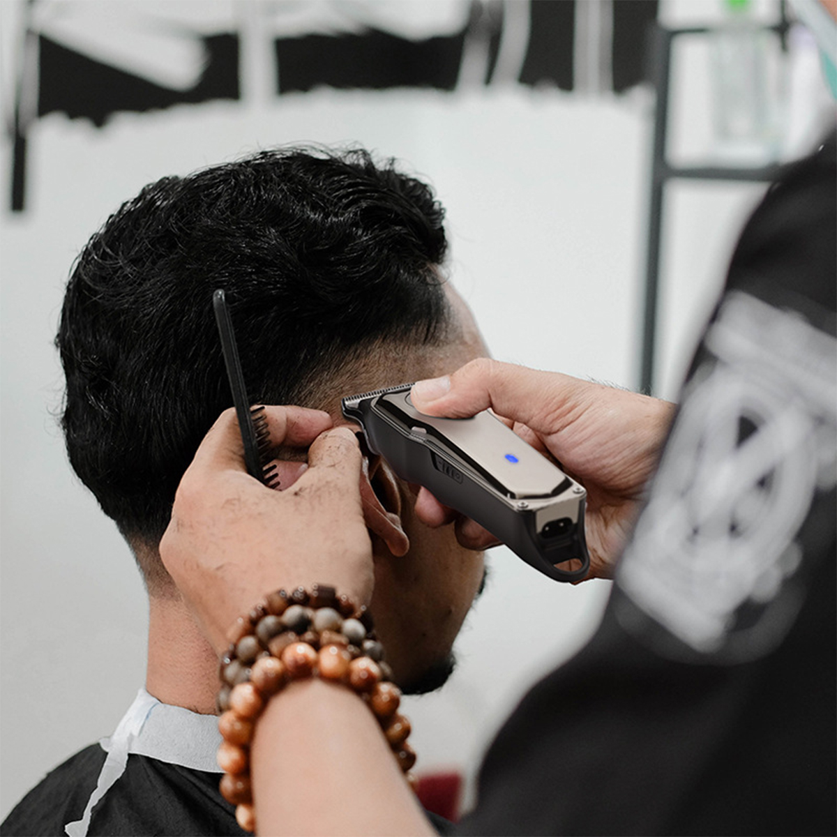 Ein Friseur schneidet einem Mann die Haare. Der Friseur benutzt einen Trimmer und einen Kamm. Der Mann trägt ein Armband.